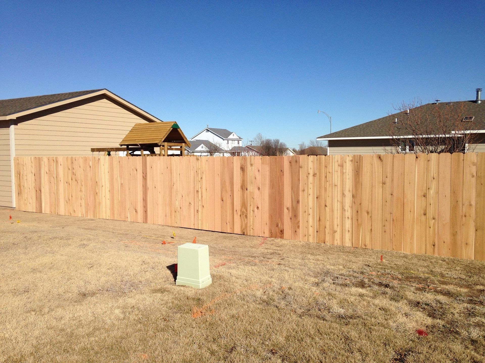 A new wooden privacy fence spans across a suburban yard on a sunny day with houses visible in the background.