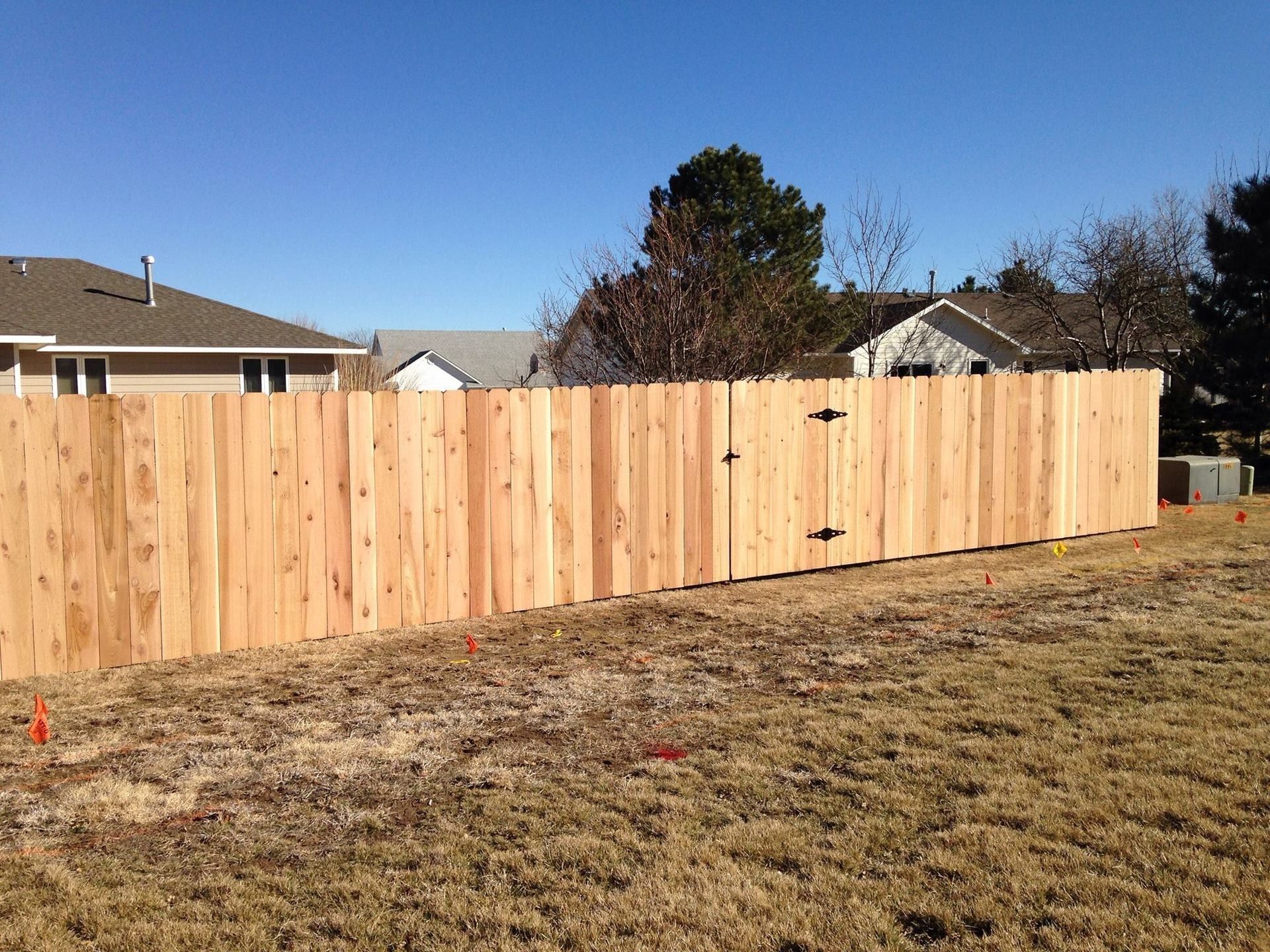 A new, tall, light-colored wooden privacy fence stands in a grassy yard on a sunny day with houses in the background.