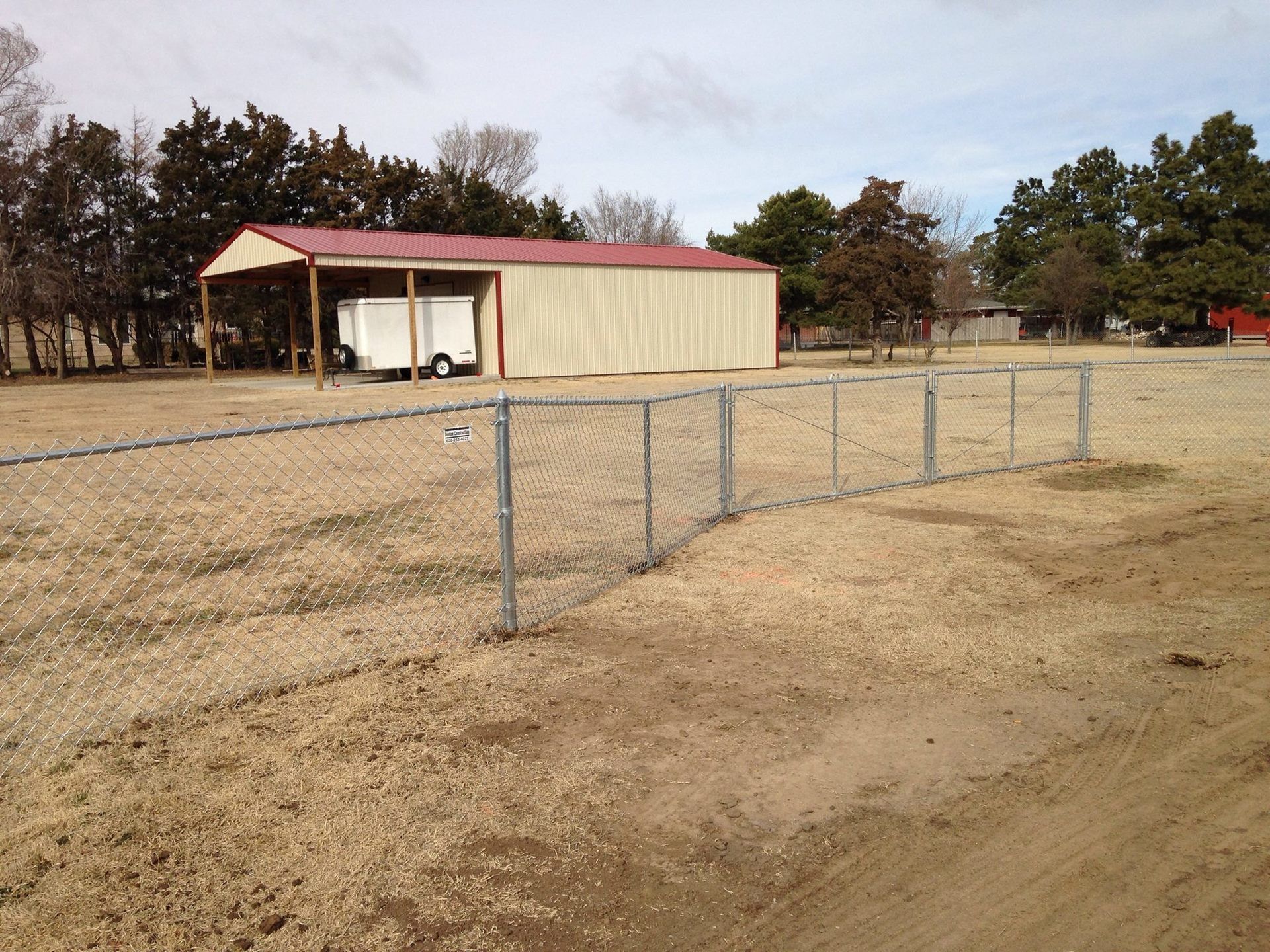 A chain-link fence sits on dry grass in front of a metal-roofed structure with a parked trailer.
