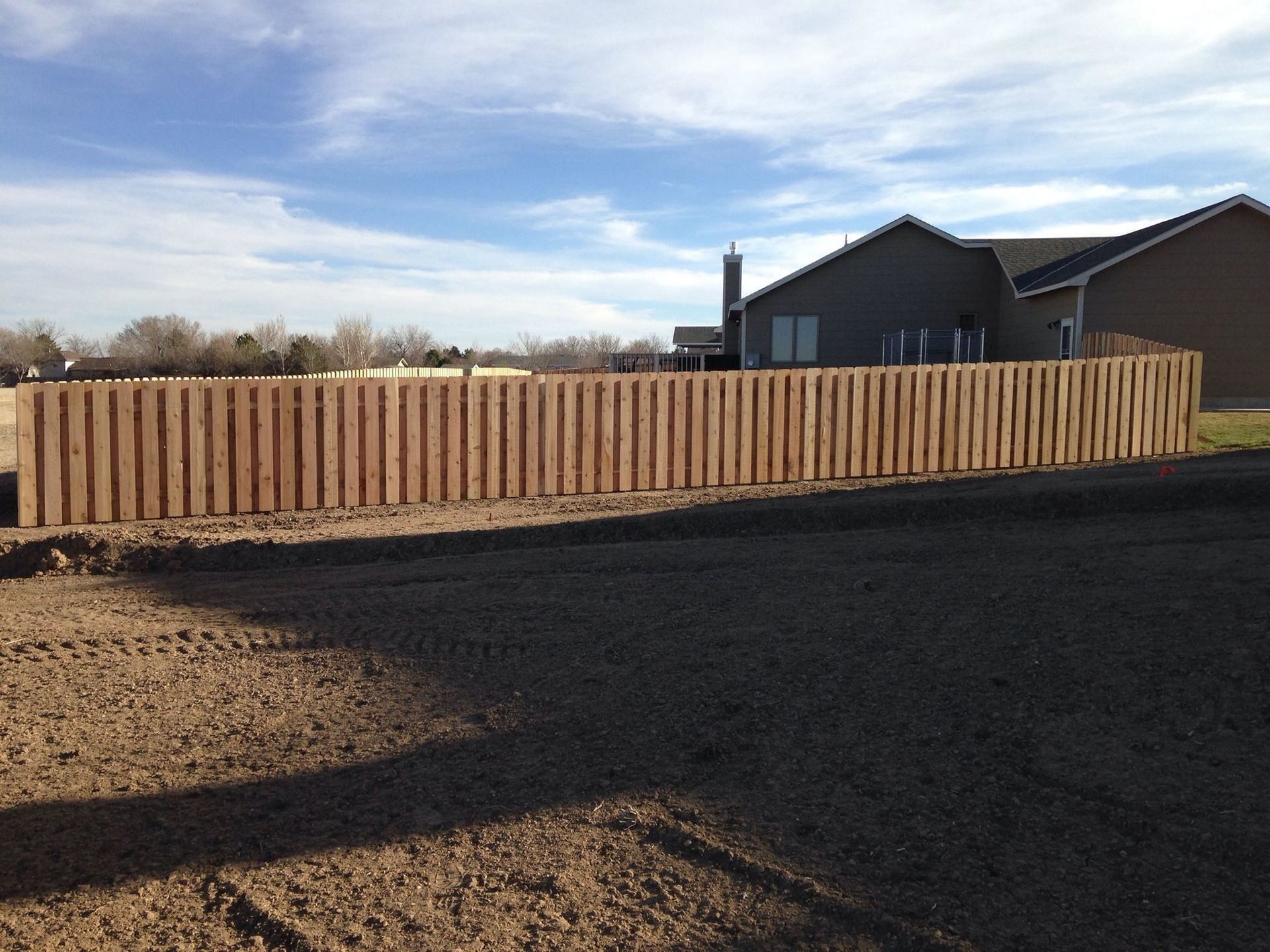 A newly installed wooden picket fence stands in front of a house against a blue sky over a dirt construction site.