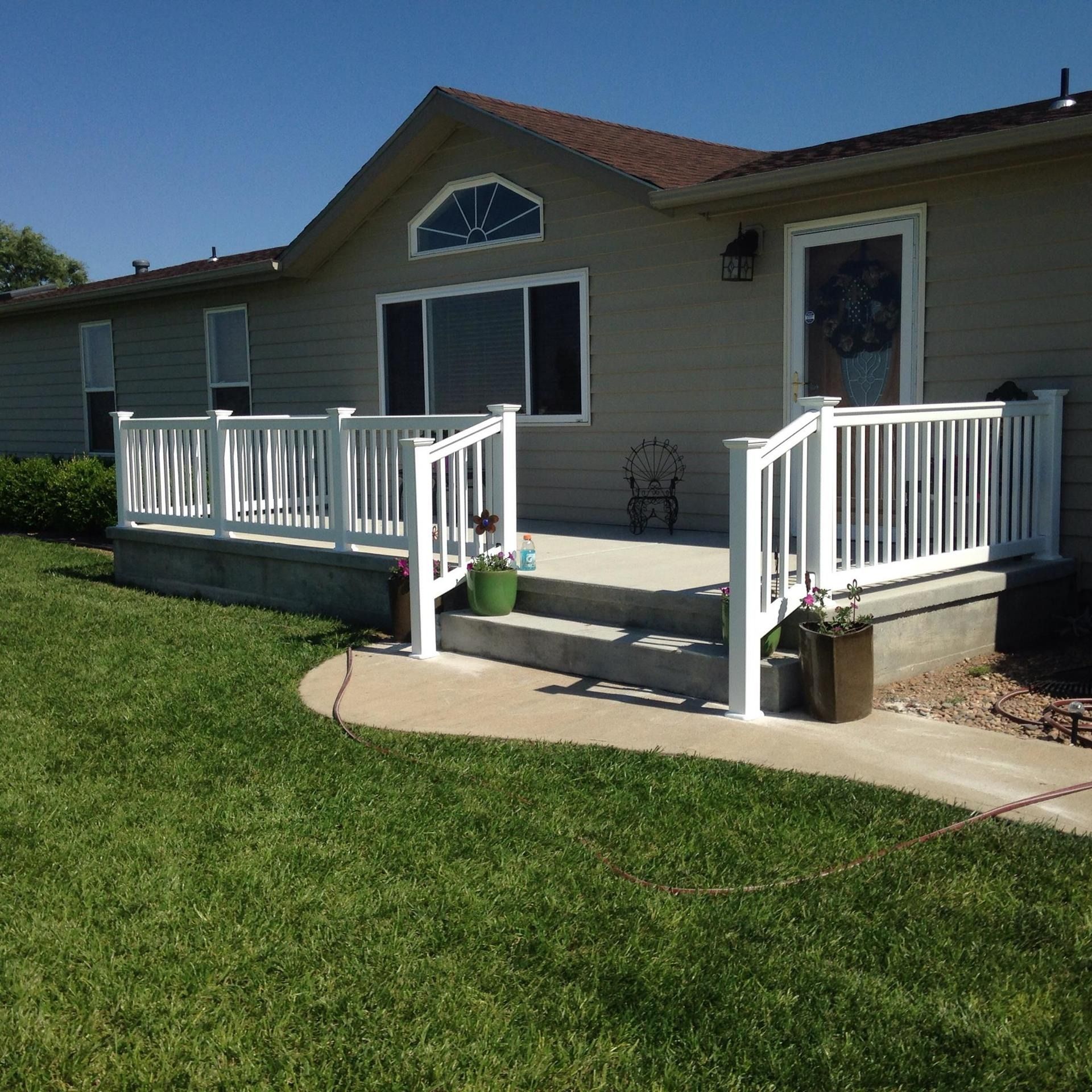 A beige house features a white deck and concrete stairs with white vinyl railings, surrounded by a green grass lawn.