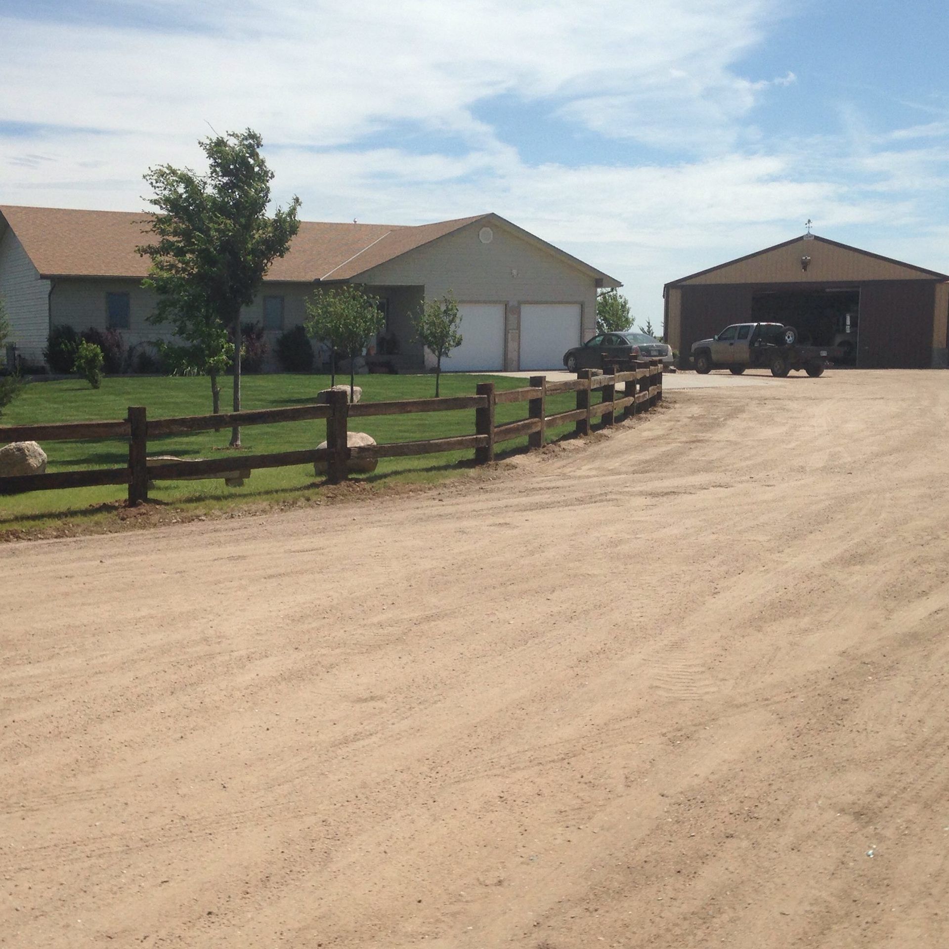 A tan house with a two-car garage and an adjacent detached workshop, separated by a wood fence from a dirt driveway.