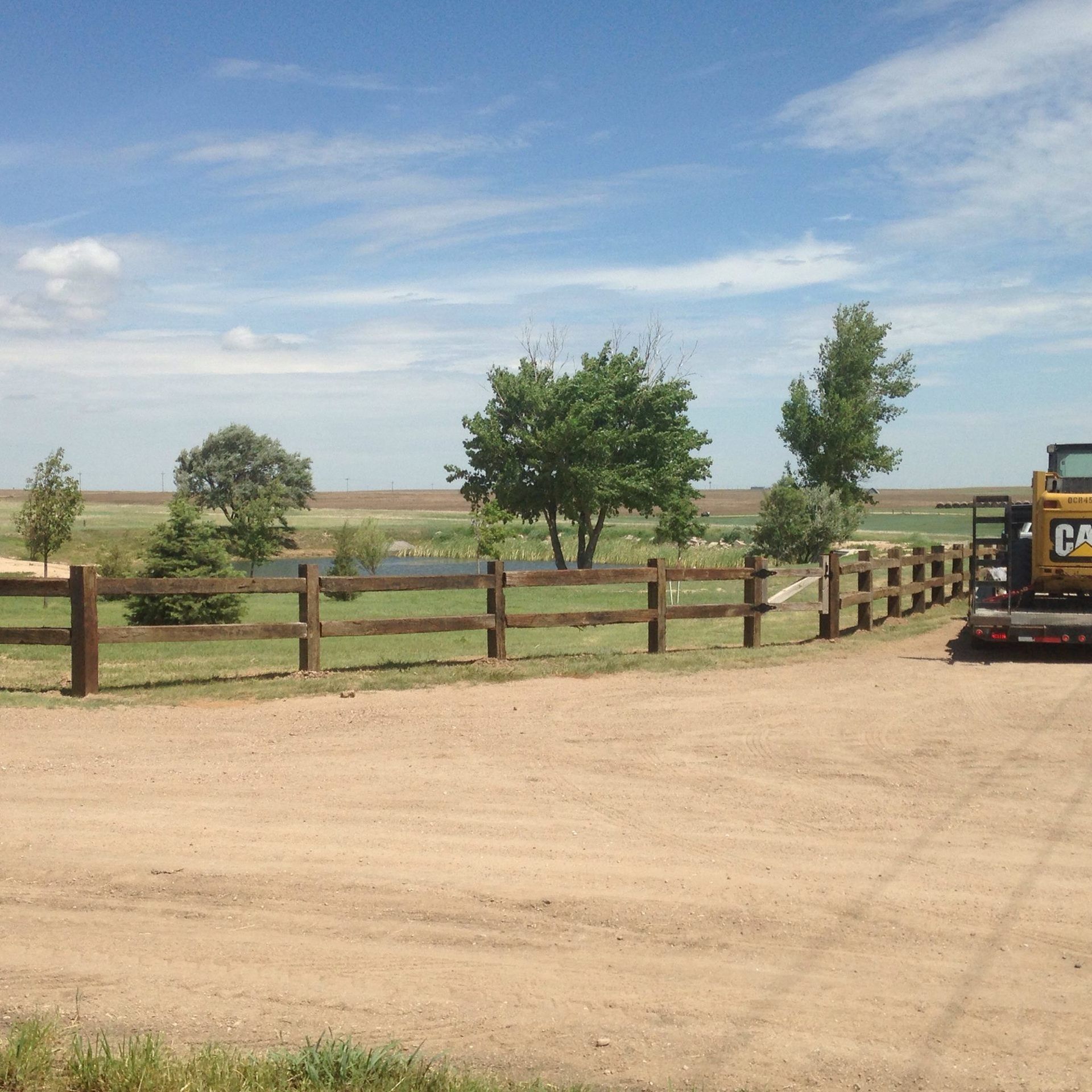A wooden fence runs through a grassy field with trees and a small pond, under a blue sky with light clouds.