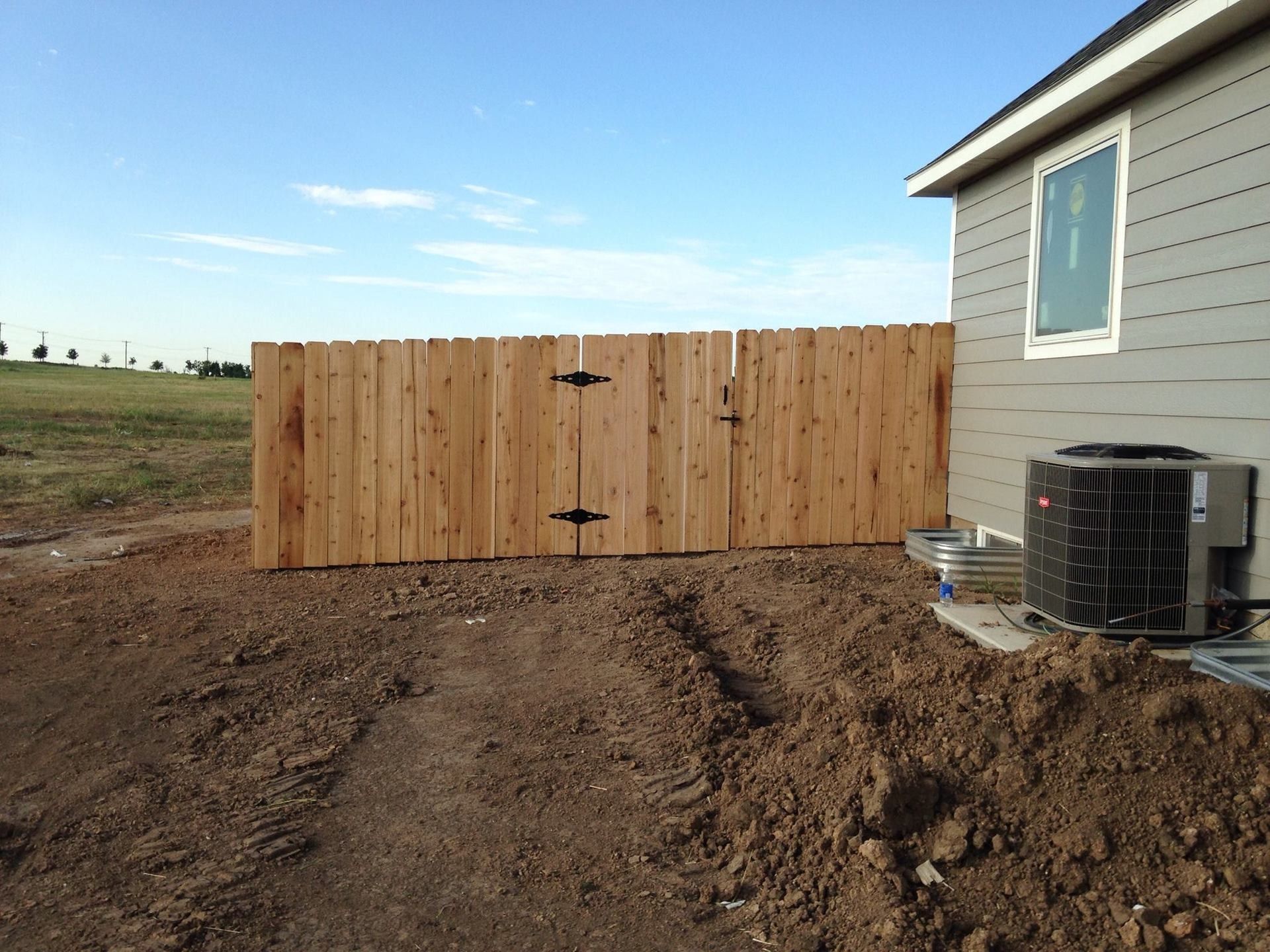 A new wooden fence stands next to a gray house, bordering an unfinished dirt yard under a clear sky.
