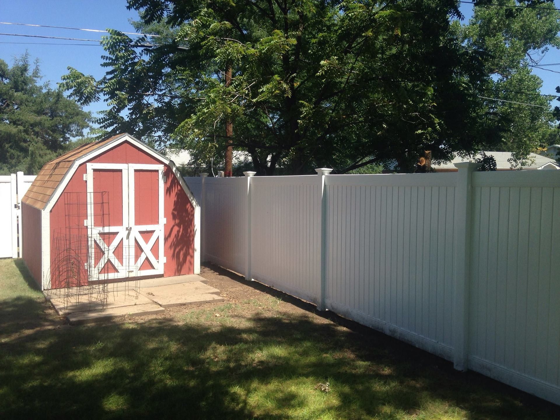A red shed with white trim sits on a stone patio in a sunny backyard next to a tall, white privacy fence.