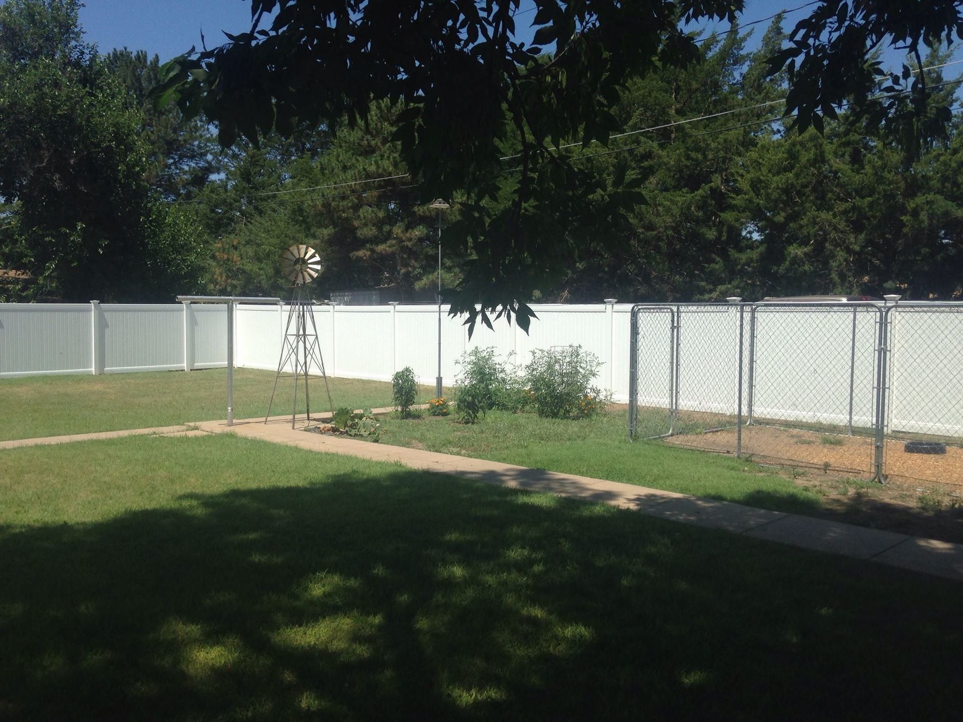 A fenced backyard with a green lawn, a stone walkway, a decorative metal windmill, and a wire dog run.