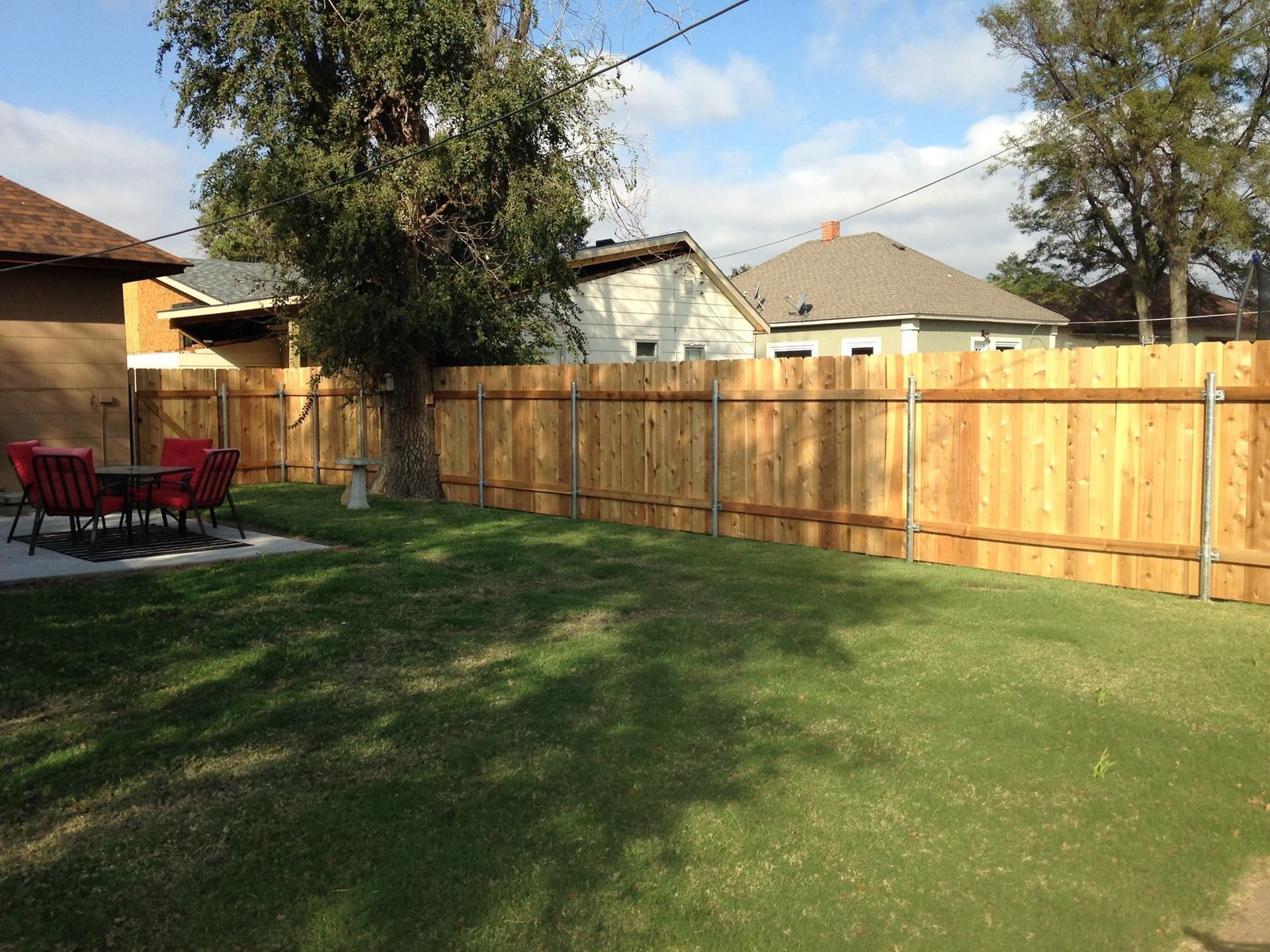 A backyard with a green lawn, red chairs on a patio, a large tree, and a wooden privacy fence under a blue sky.