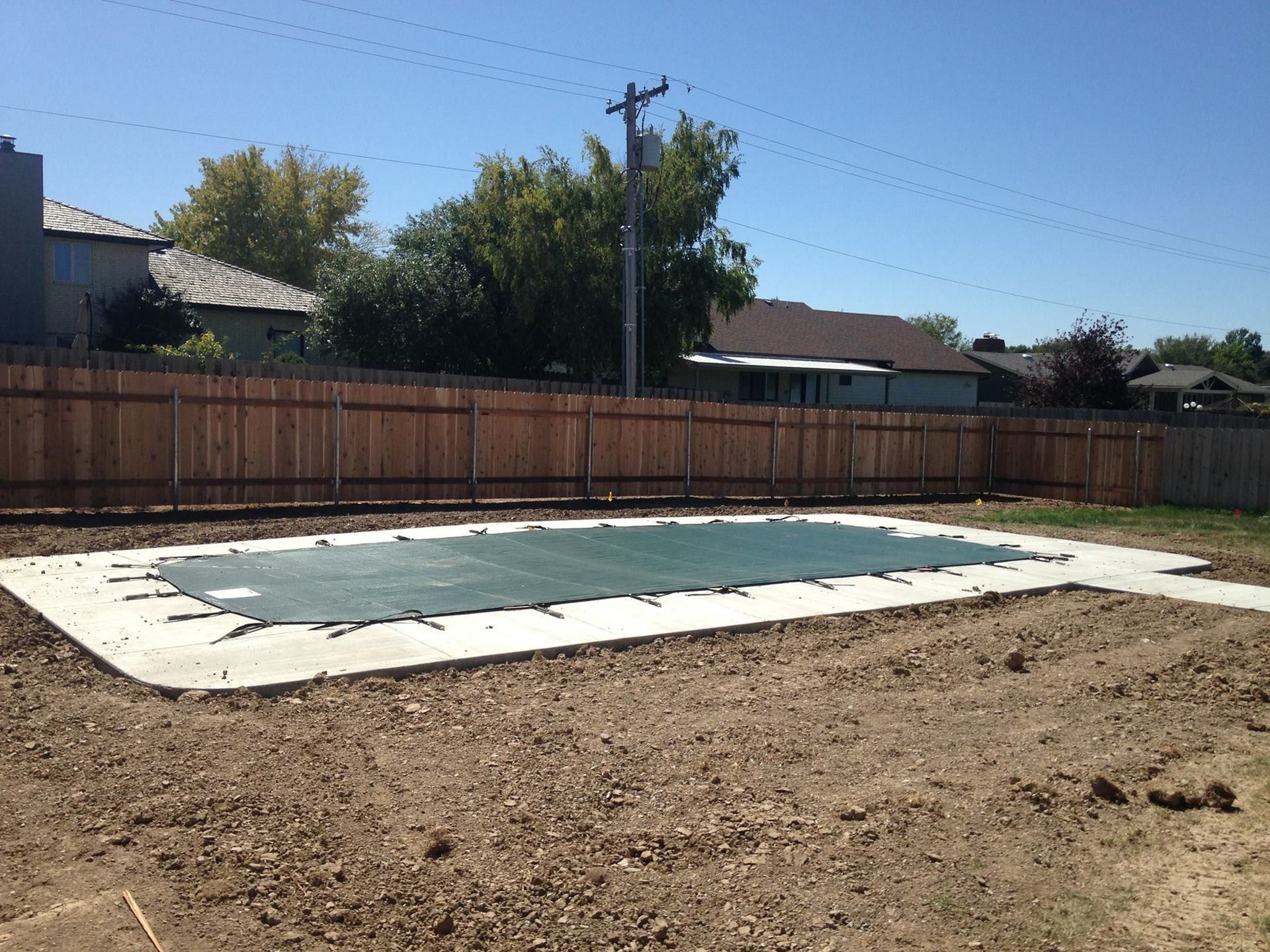 A rectangular swimming pool covered by a green mesh safety tarp, set in a backyard with a wooden fence and dirt yard.