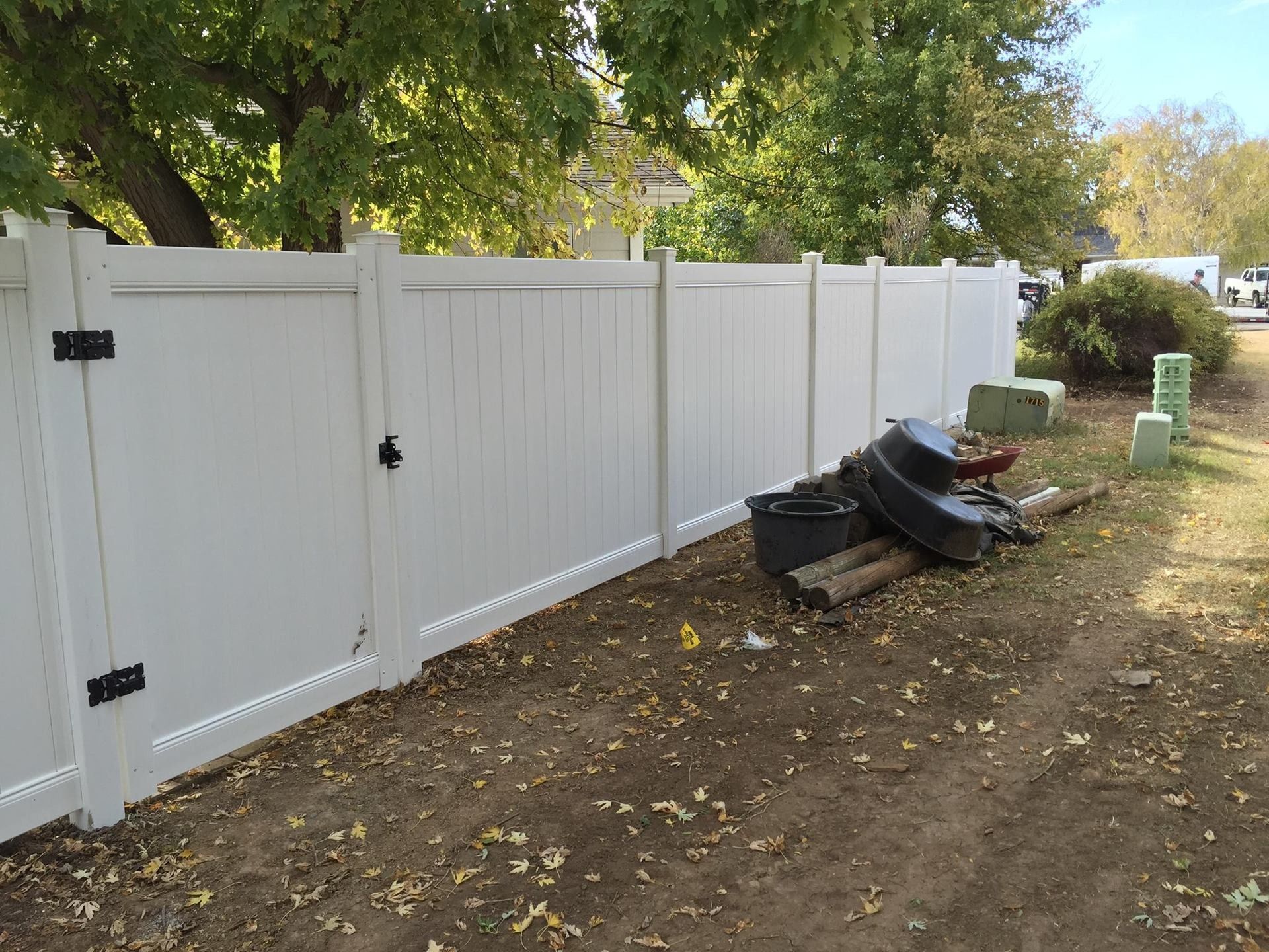 A white vinyl privacy fence with a gate stands along a dirt ground area near trees and some yard debris.