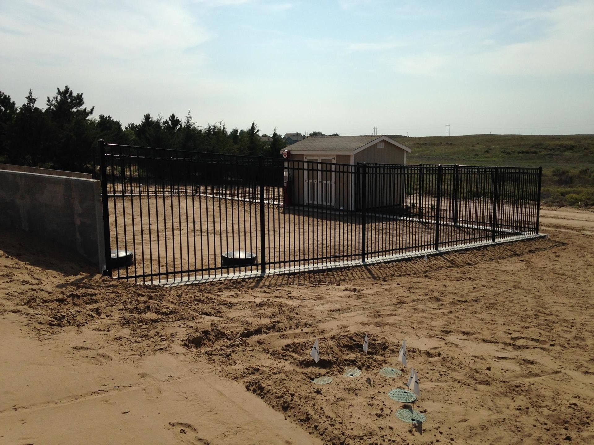A small beige shed stands inside a black metal fence on a dirt lot, near a concrete wall and some pine trees.