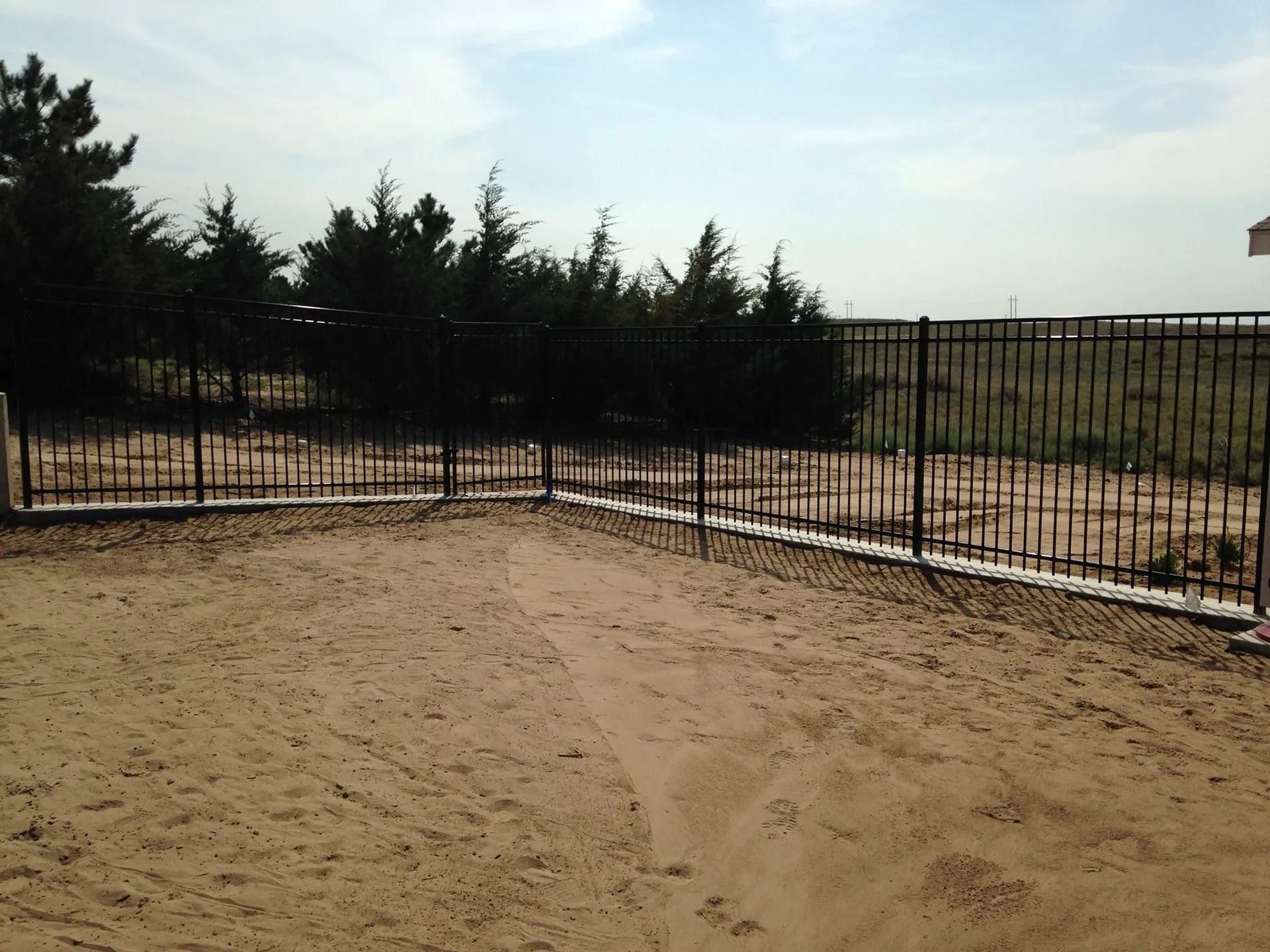 A black metal fence surrounds a sandy yard with evergreen trees in the background under a blue sky.