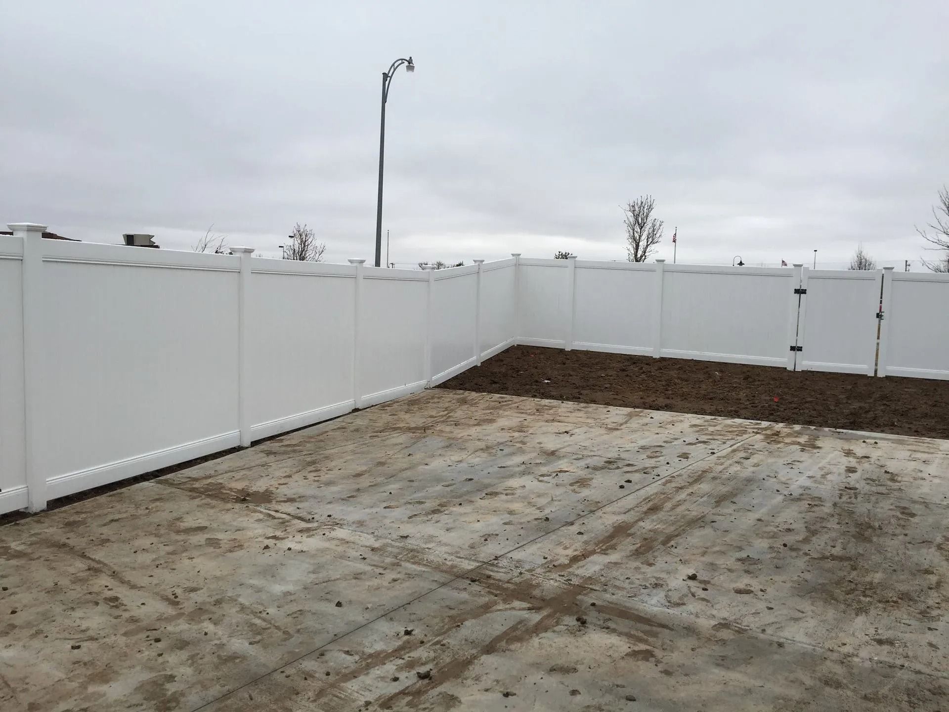 A white vinyl fence borders a dirt backyard under an overcast sky.
