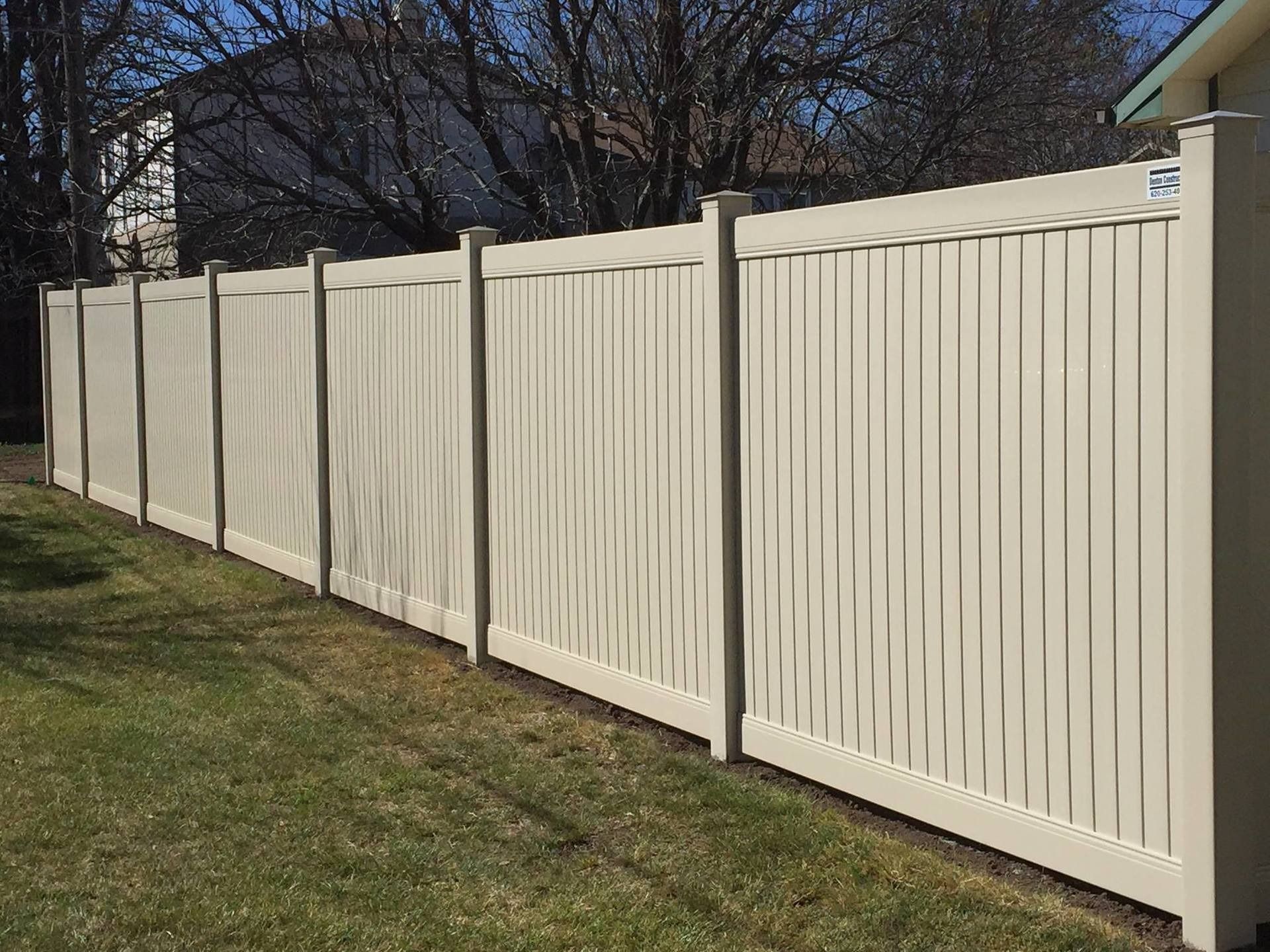 A long, solid beige vinyl privacy fence stands on a grassy lawn under a clear blue sky.