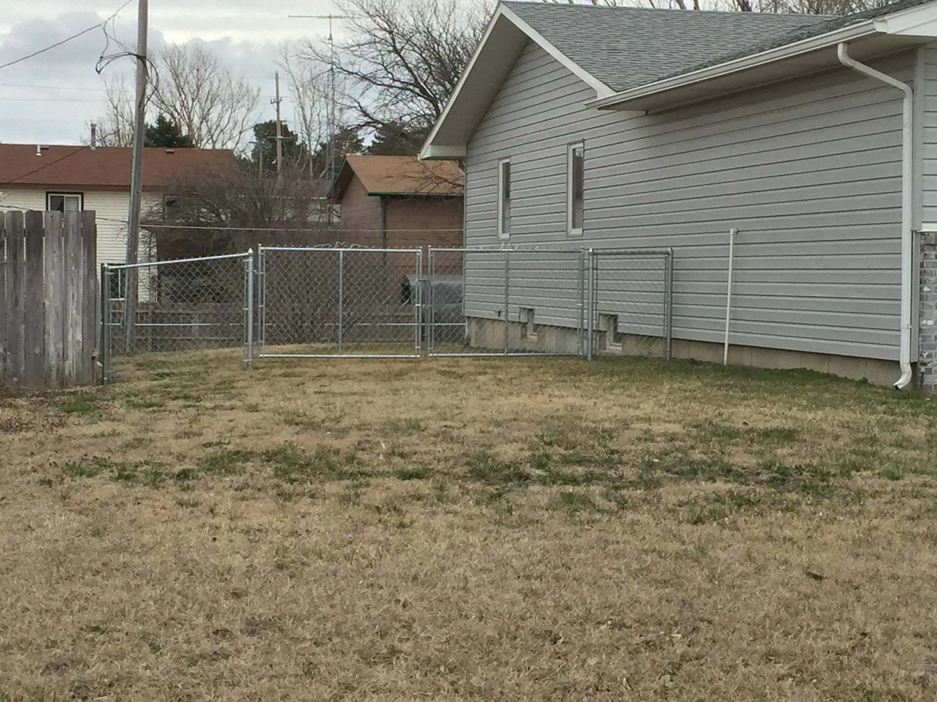 A chain-link fence divides a yard next to a gray-sided house under a cloudy sky.