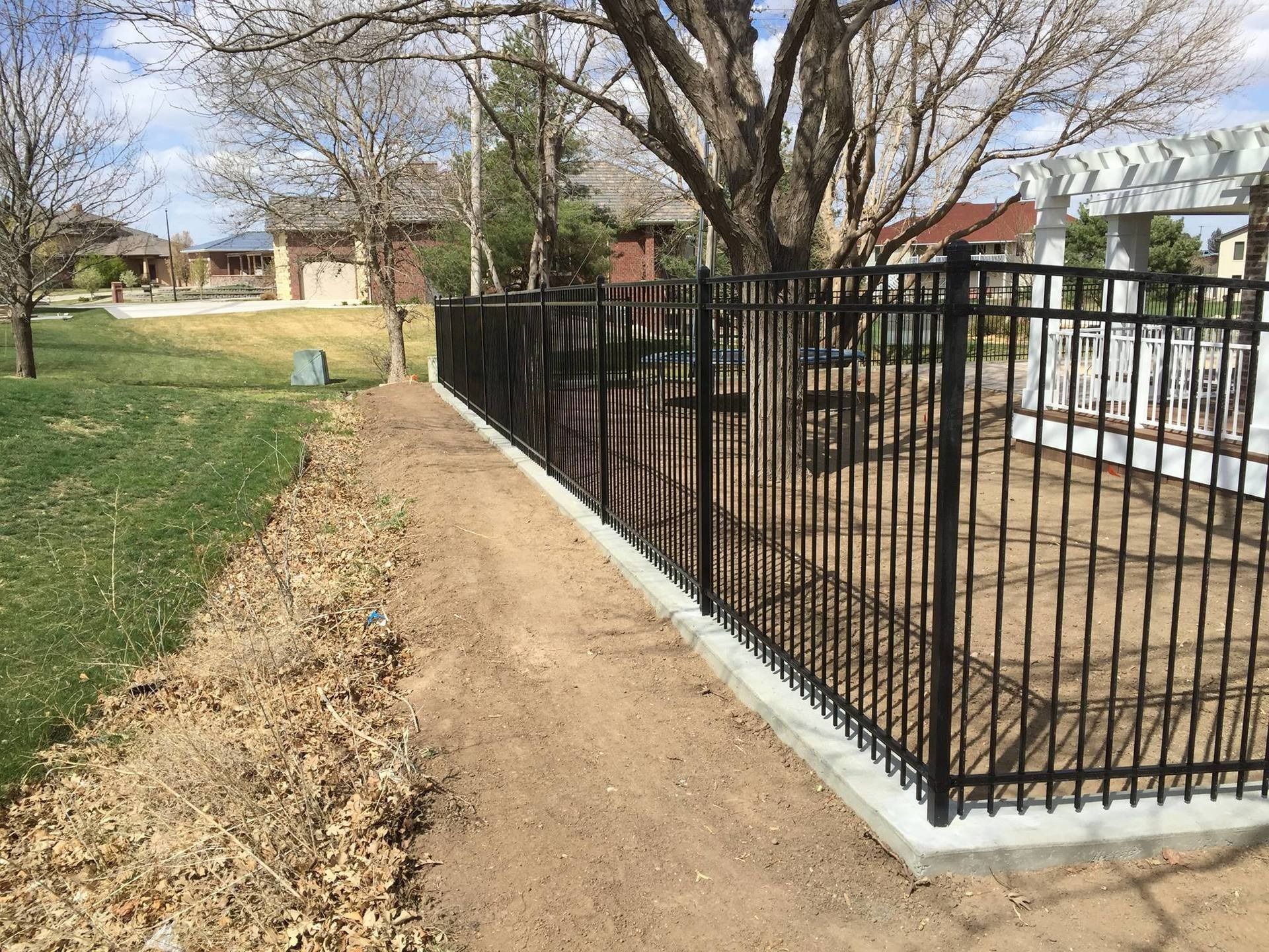 A black metal fence installed on a concrete curb runs along a dirt path beside a grassy yard with trees and a pergola.