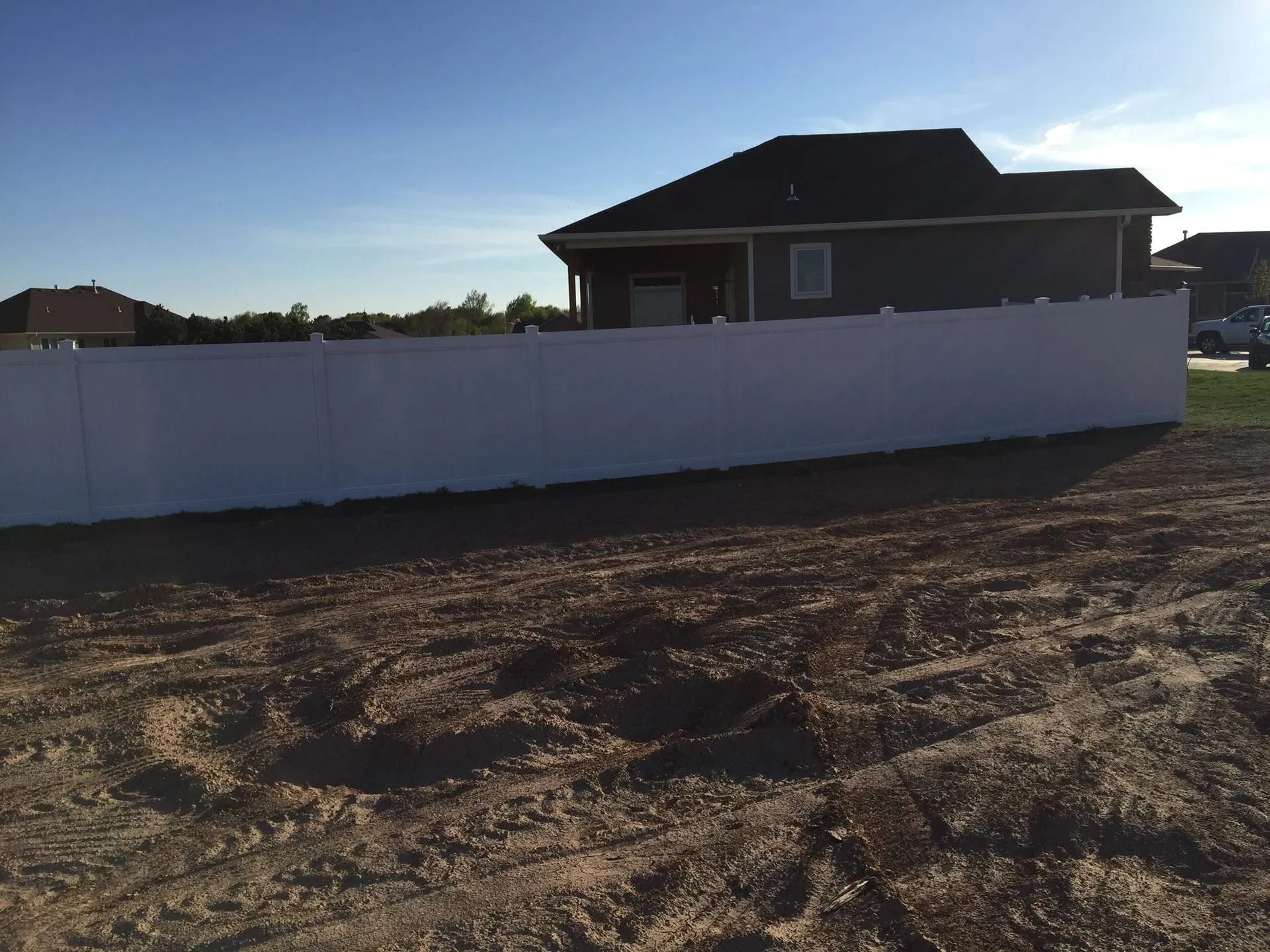 A long white privacy fence stands in a dirt yard in front of a dark-roofed residential house under a clear blue sky.