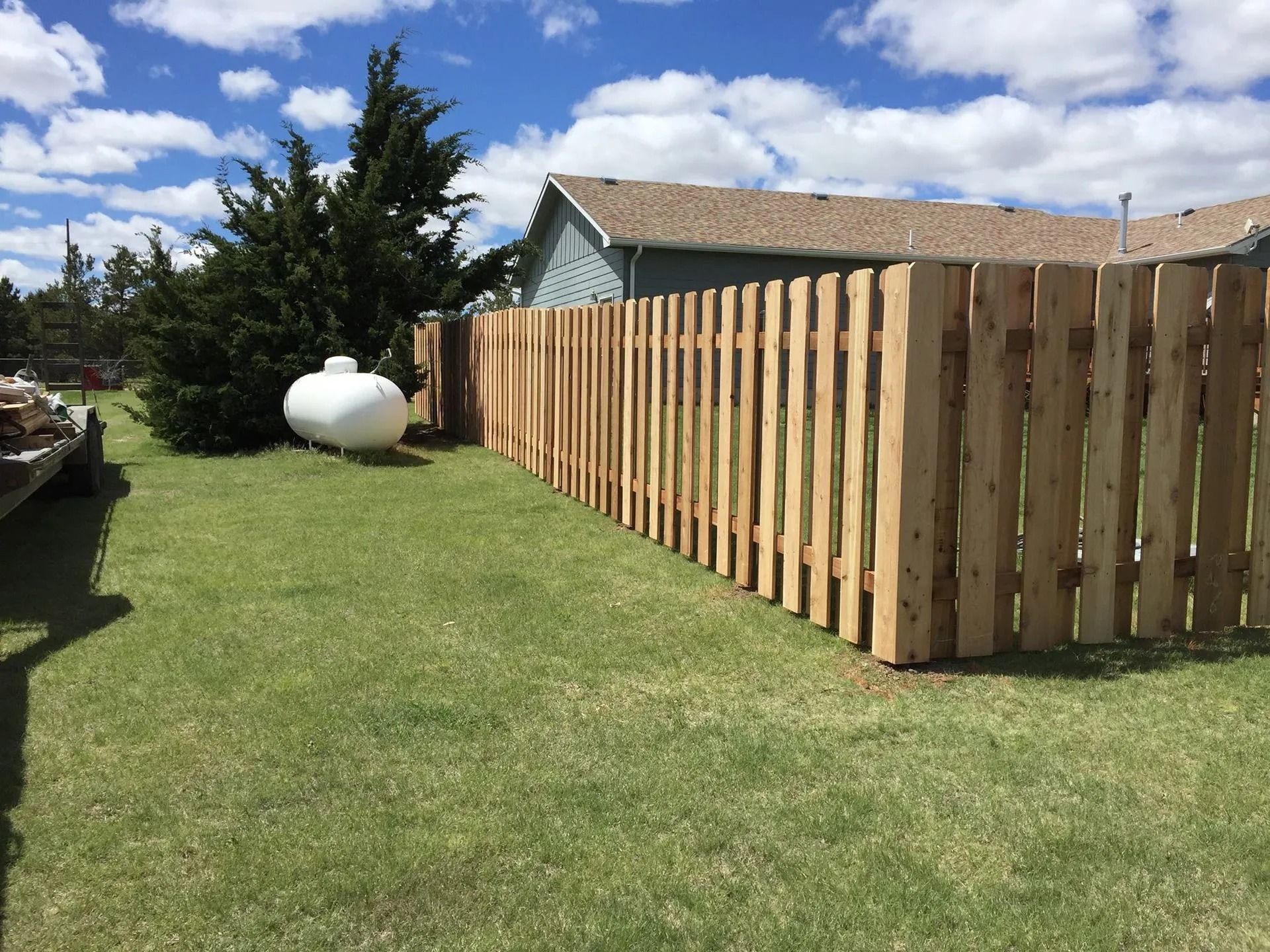 A newly installed natural wood picket fence stands in a grassy yard next to a propane tank and a tree under a blue sky.