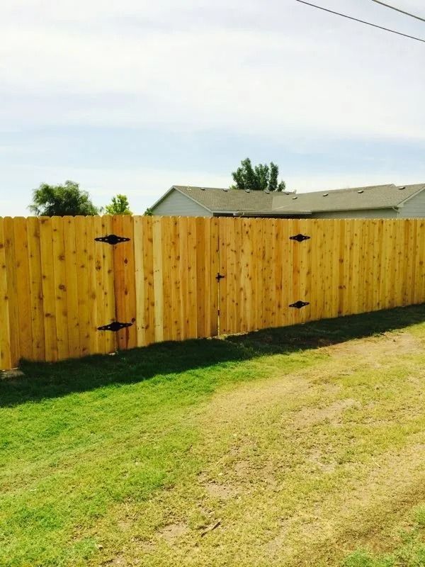 A tall, stained-wood fence with a gate and black hardware stands in a grassy yard under a partly cloudy sky.