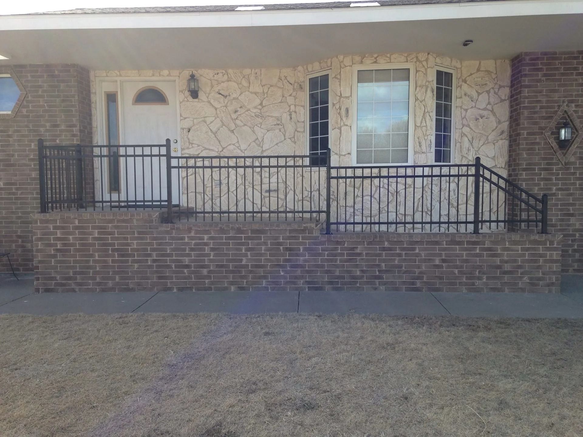 A brick porch with a black metal railing leads to a white front door and a window on a light-colored stone facade.