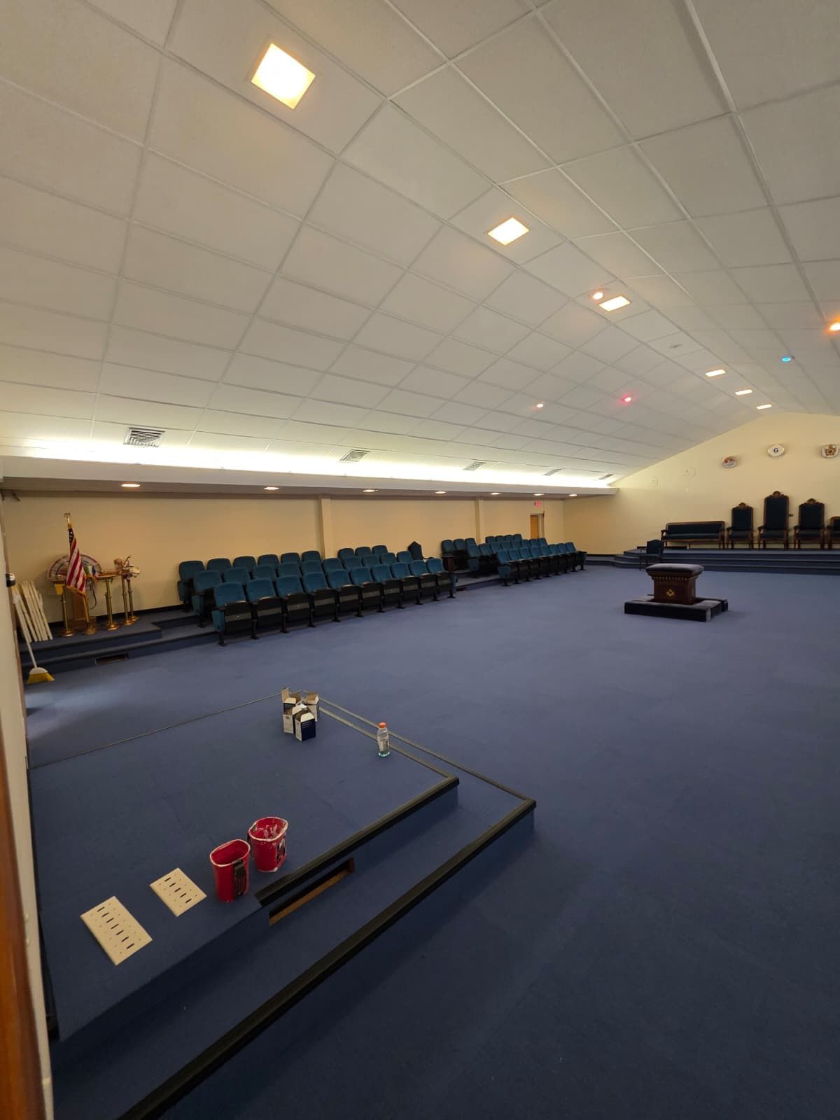 Empty church sanctuary with blue carpet, rows of teal chairs, flags, and a pulpit stage