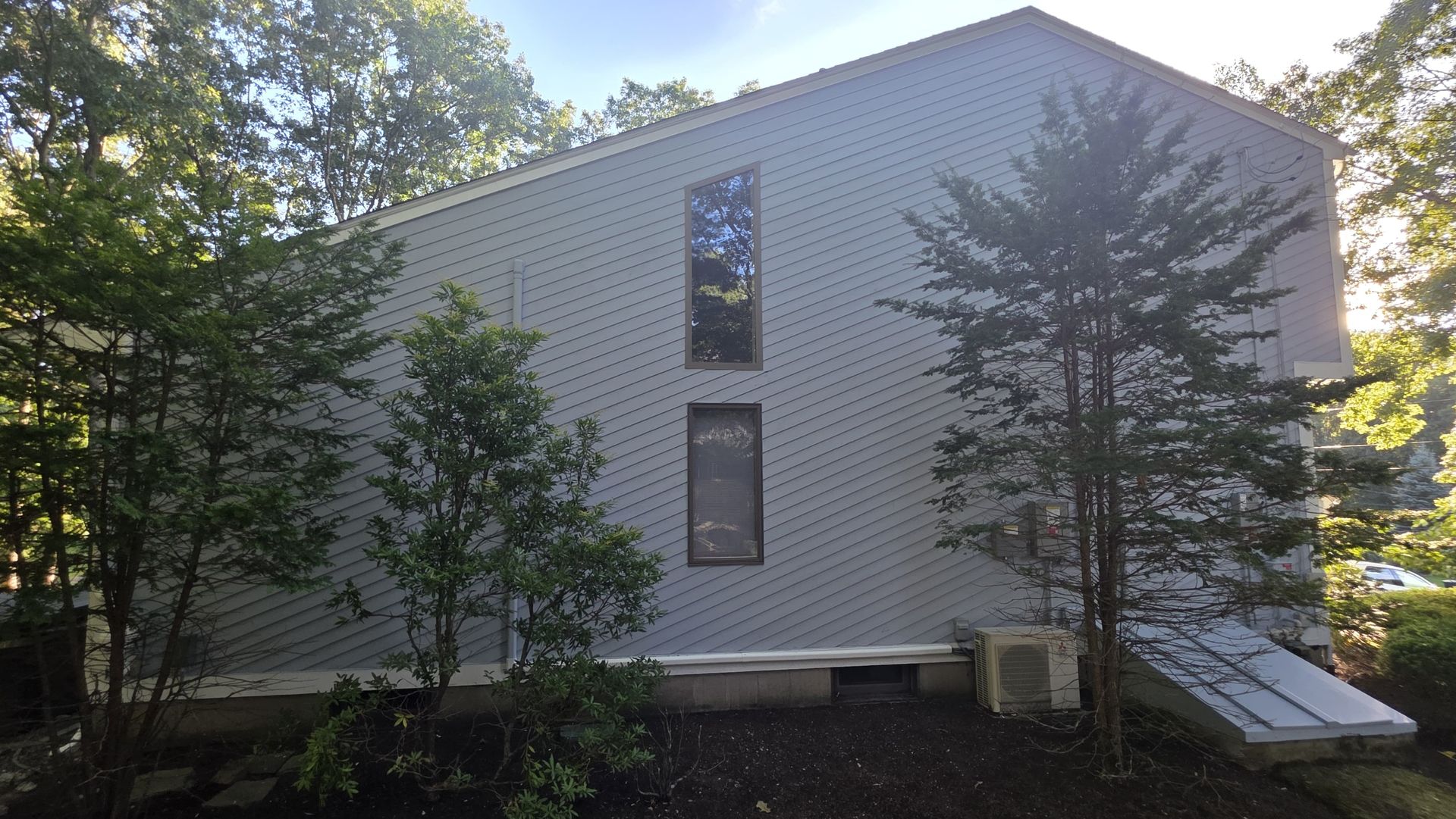 White two-story house with small windows, surrounded by trees and shaded by sunlight