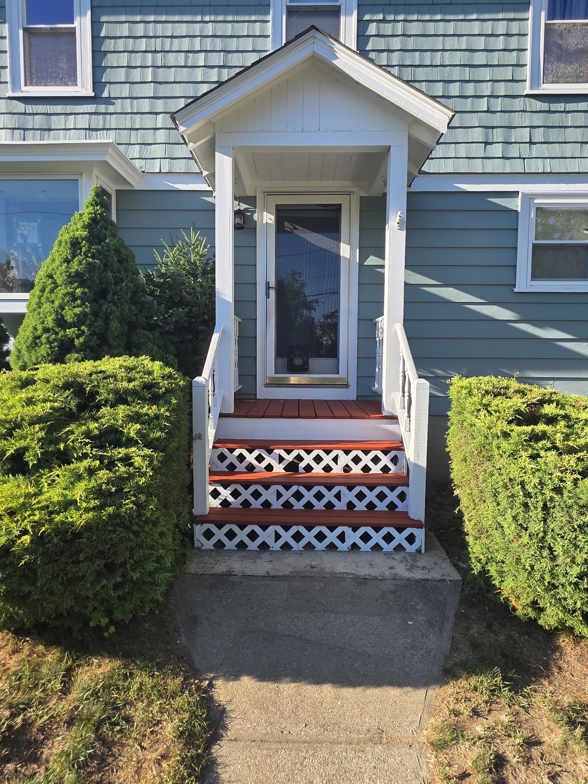 Front entrance of a blue house with white trim, red-and-white steps, and neatly trimmed bushes