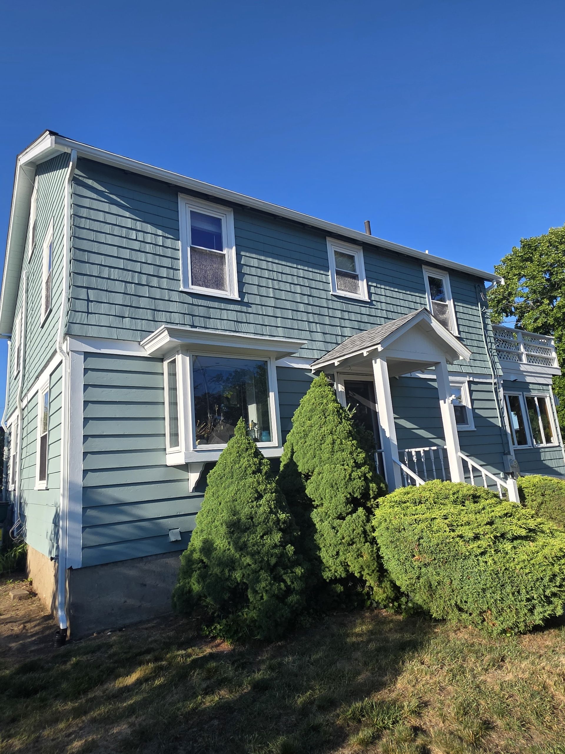 Blue two-story house with white trim, front porch, and green shrubs under a clear sky
