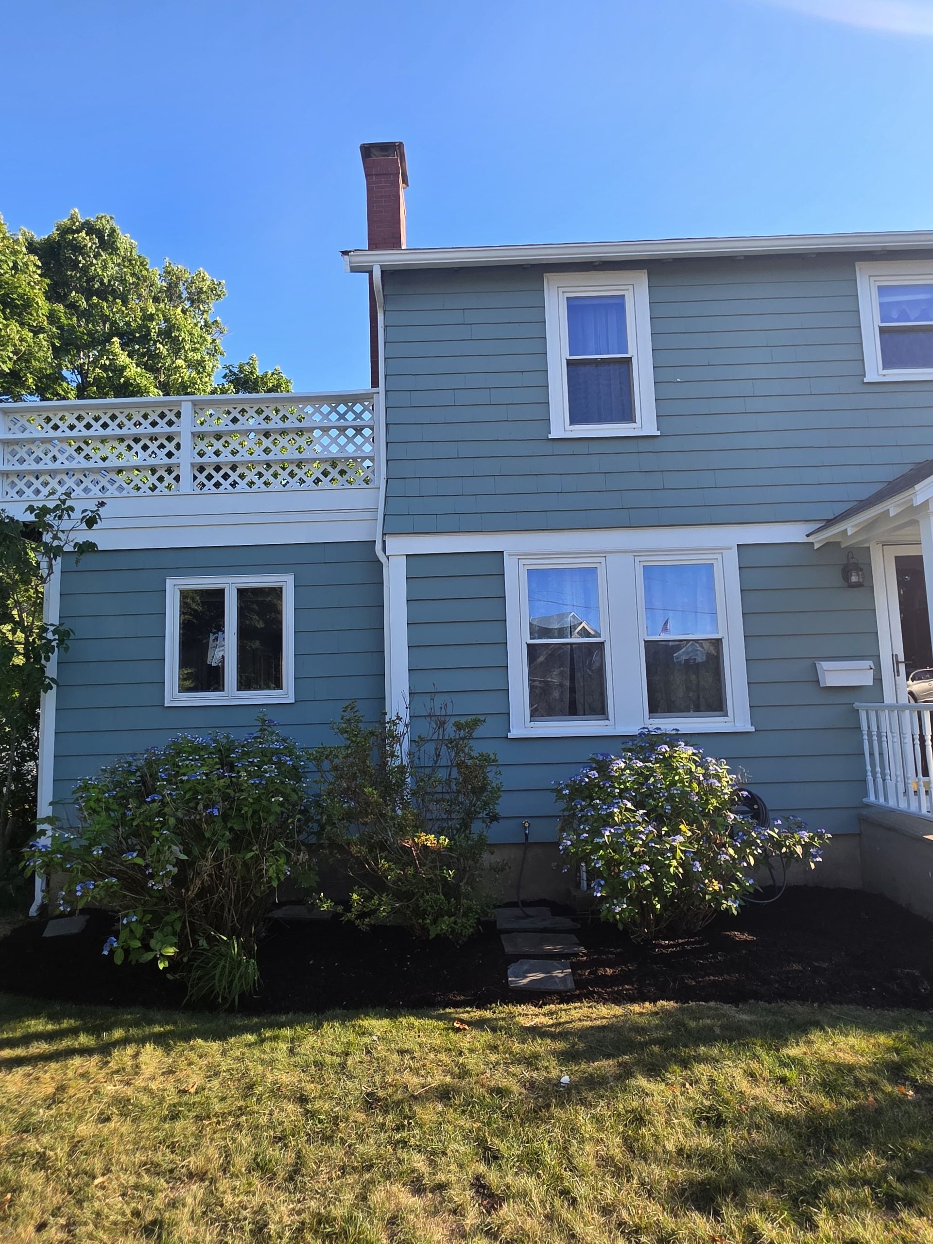 Blue two-story house with white trim and bushes in the front yard under a clear sky
