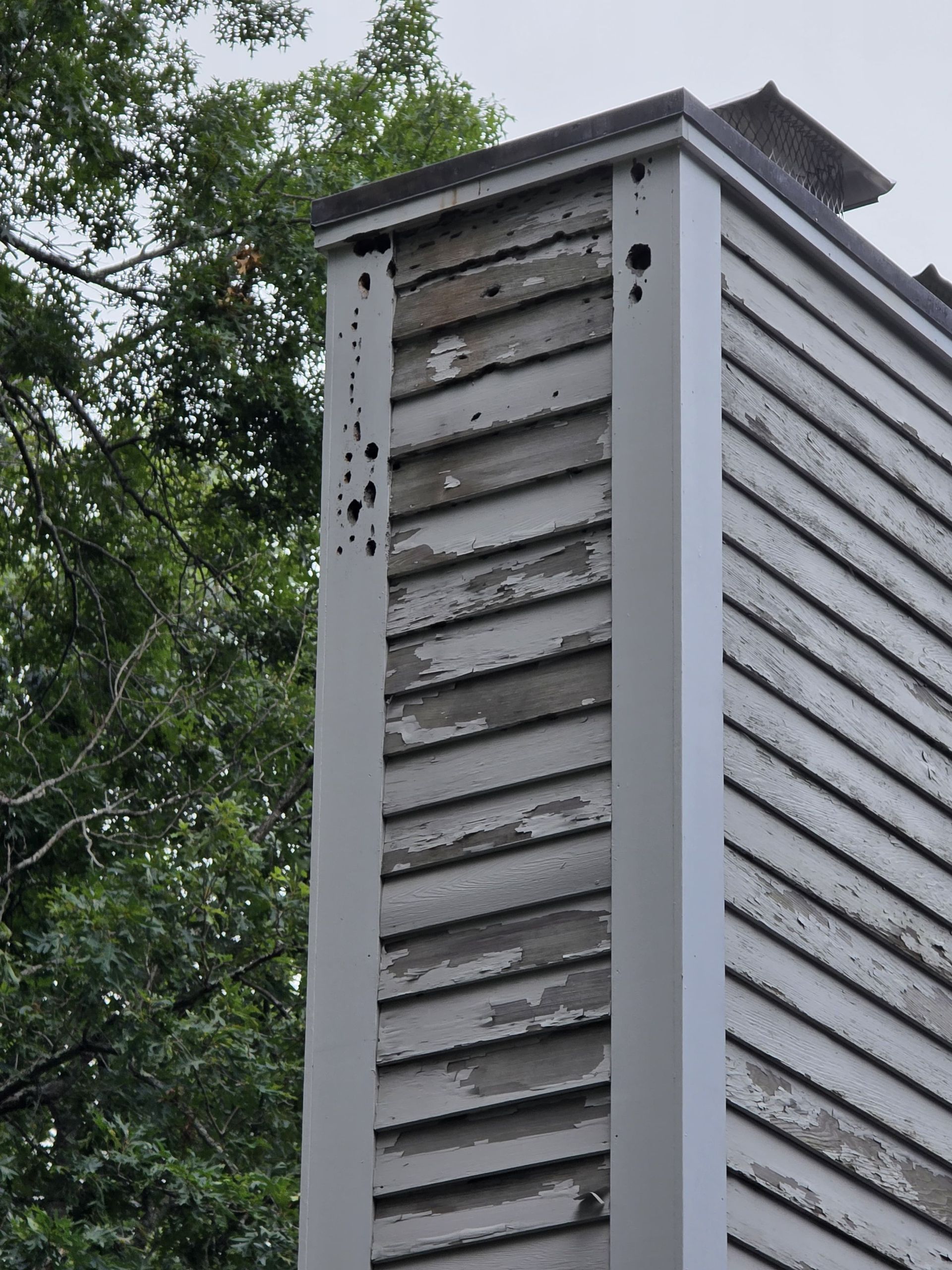 Close-up of a gray metal chimney vent with horizontal slats against leafy trees