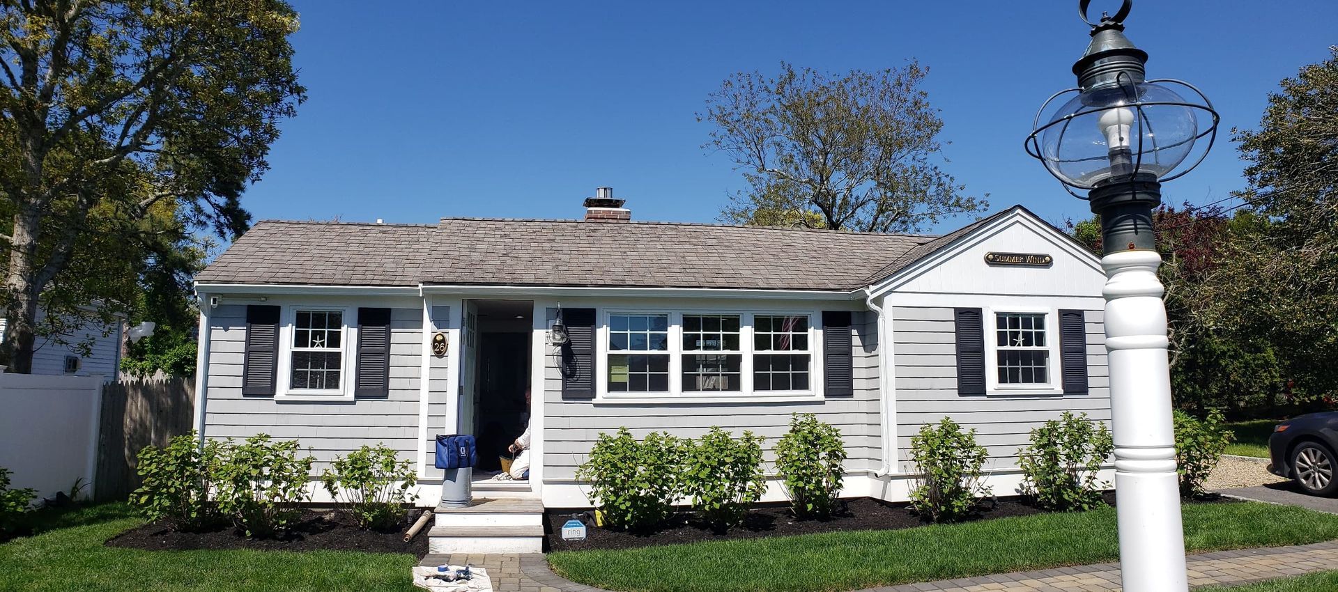 Small white house with black shutters, front steps, and a white lamp post in a sunny yard