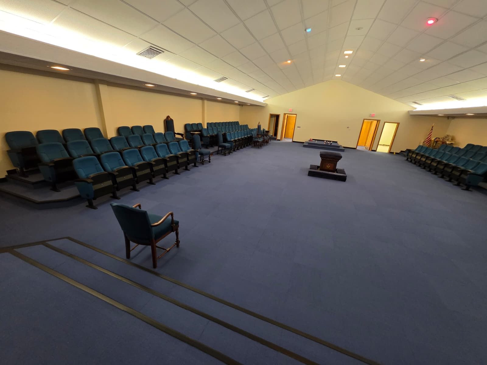 Empty auditorium with blue carpet, rows of teal seats, and a central aisle leading to lit doors.