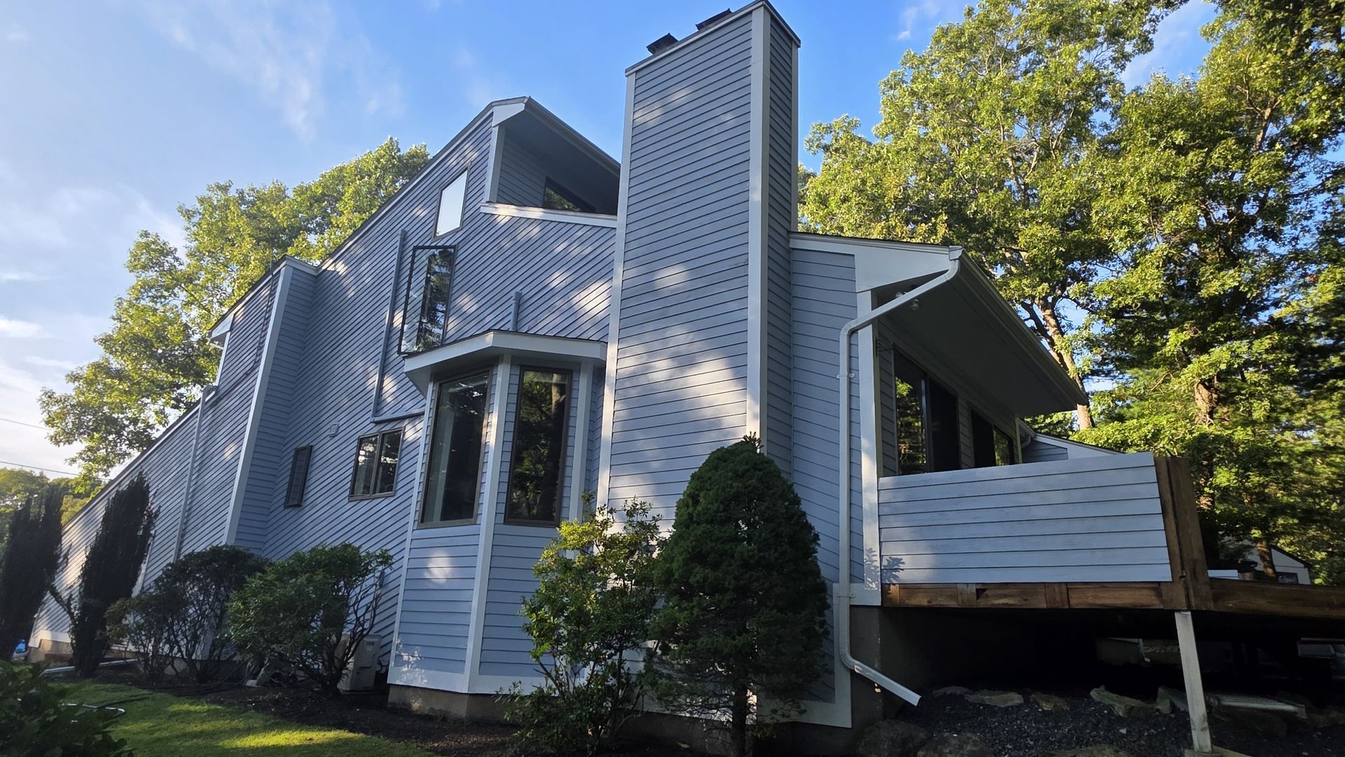 White two-story house with a raised deck and tall vertical siding, surrounded by trees in daylight