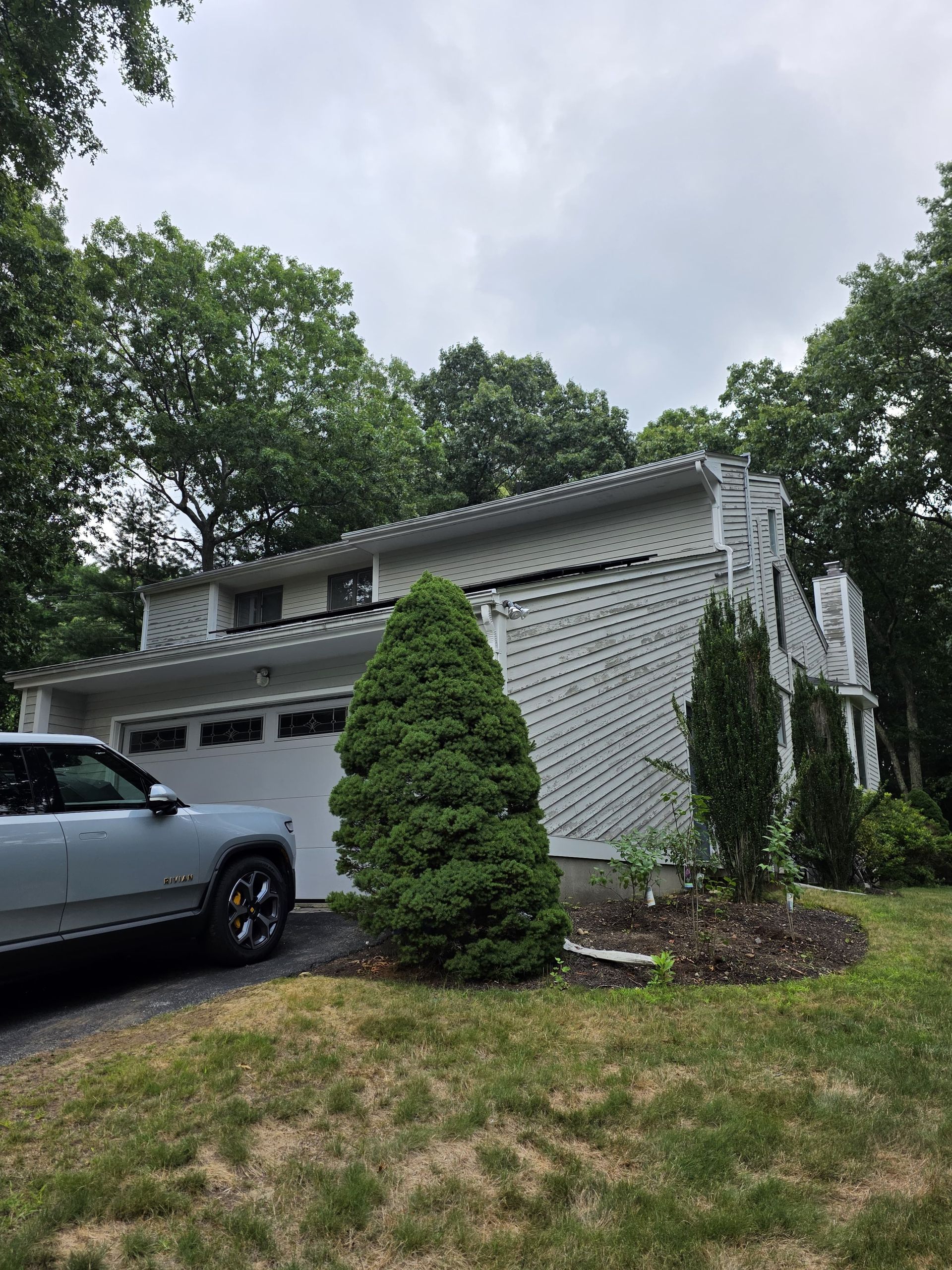 Gray SUV in front of a white house with a garage, shrubs, and trees under a cloudy sky