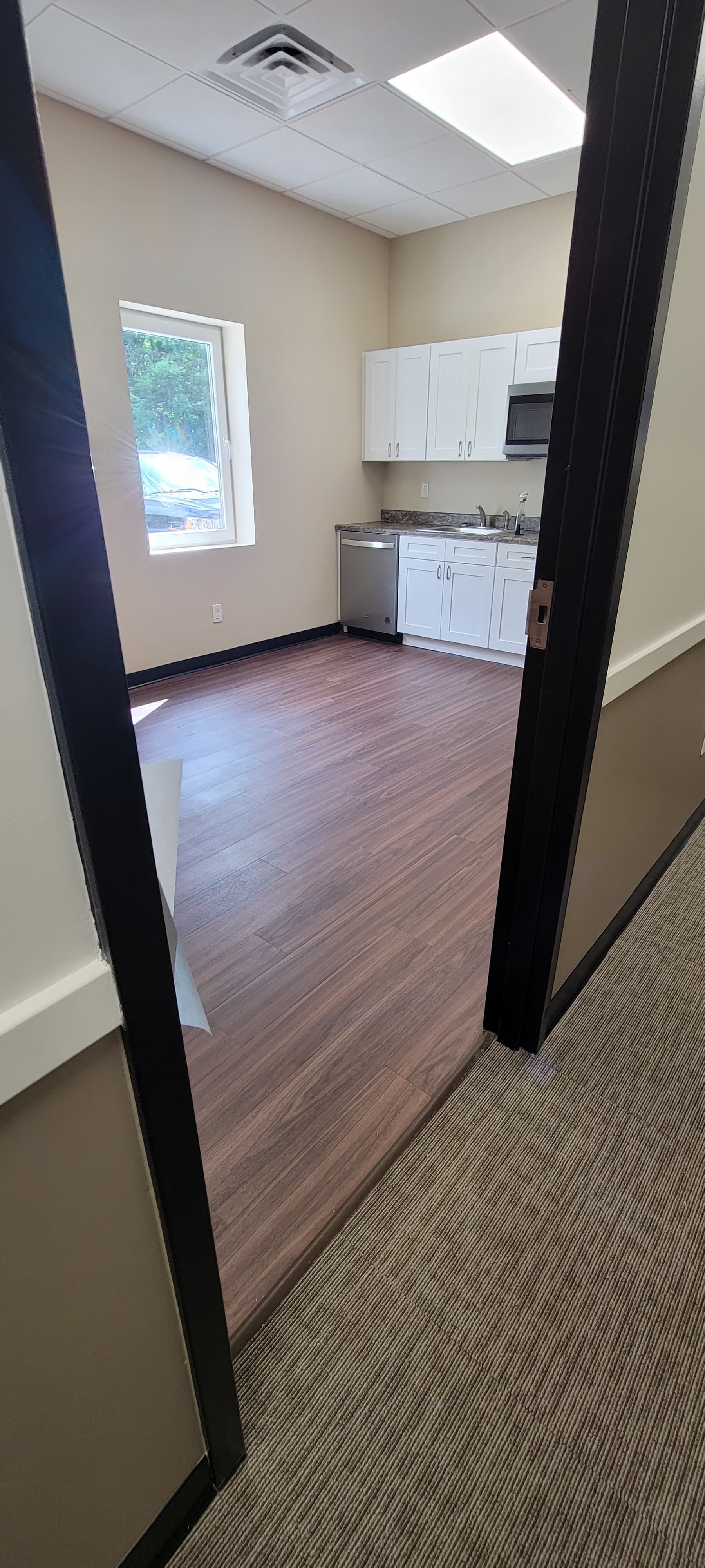 Small empty kitchenette with white cabinets, window, and wood-look floor viewed through a doorway
