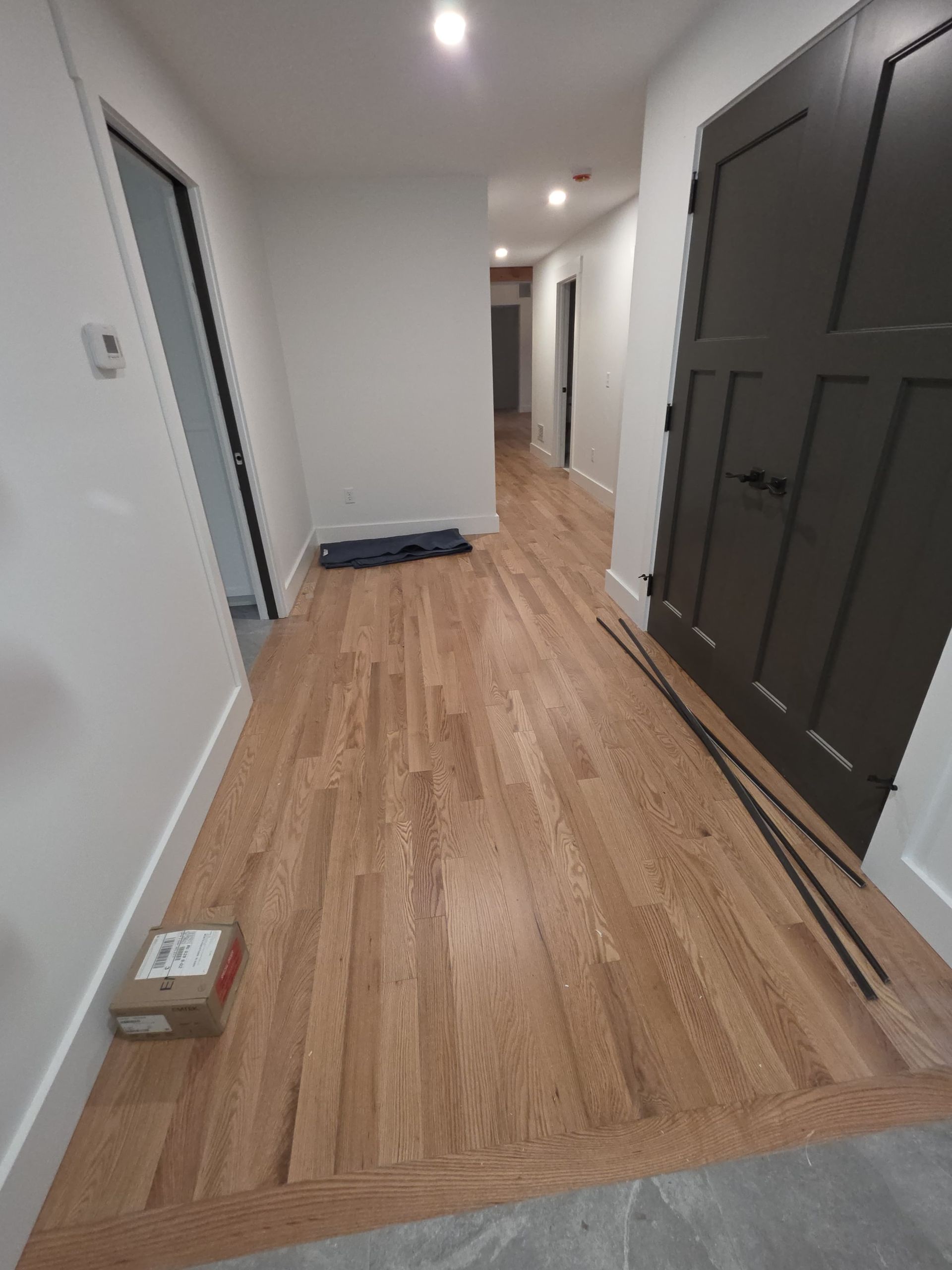 Narrow hallway with wood floor, white walls, dark front door, and recessed ceiling lights.