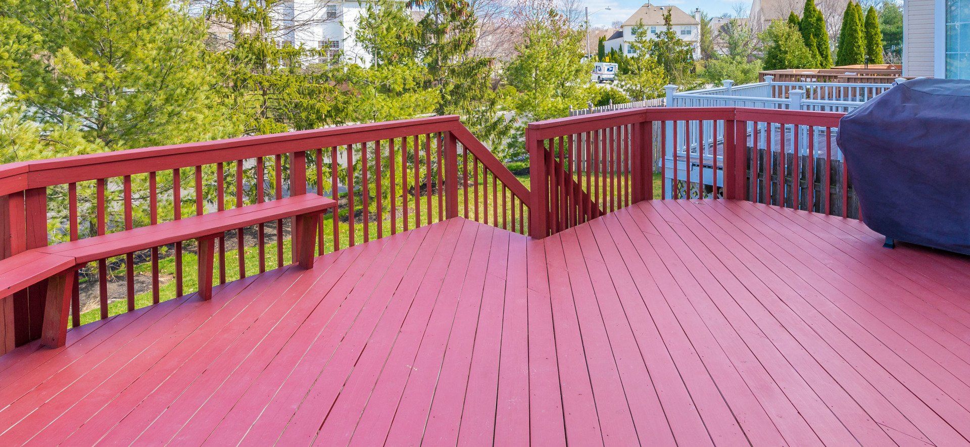 Red wooden deck with railing and bench, overlooking a backyard with trees.