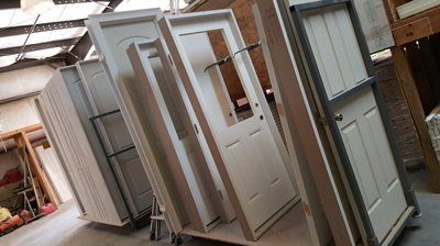 White doors leaning against a wall in a workshop; some have windows, others are paneled.