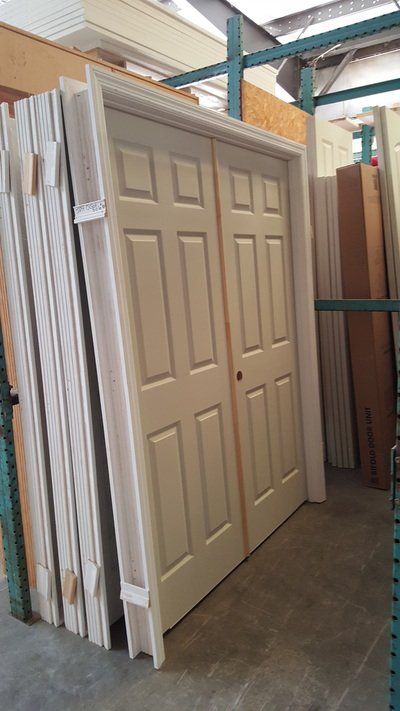 Two white interior doors with six panels each, leaning against a stack of door frames in a warehouse.