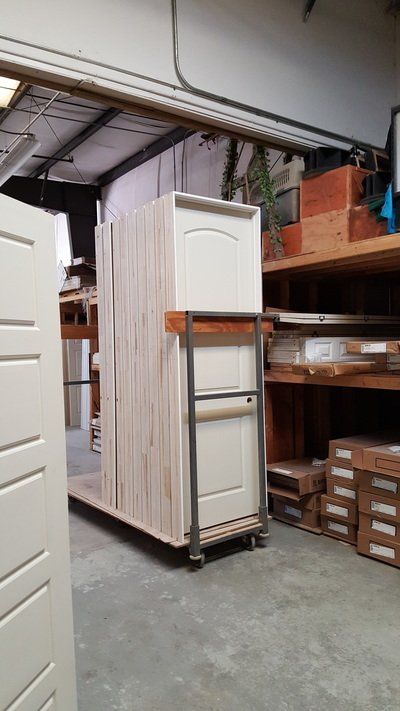 Doors stacked on a cart inside a warehouse. Beige doors are grouped together near shelving with various boxes and wood.