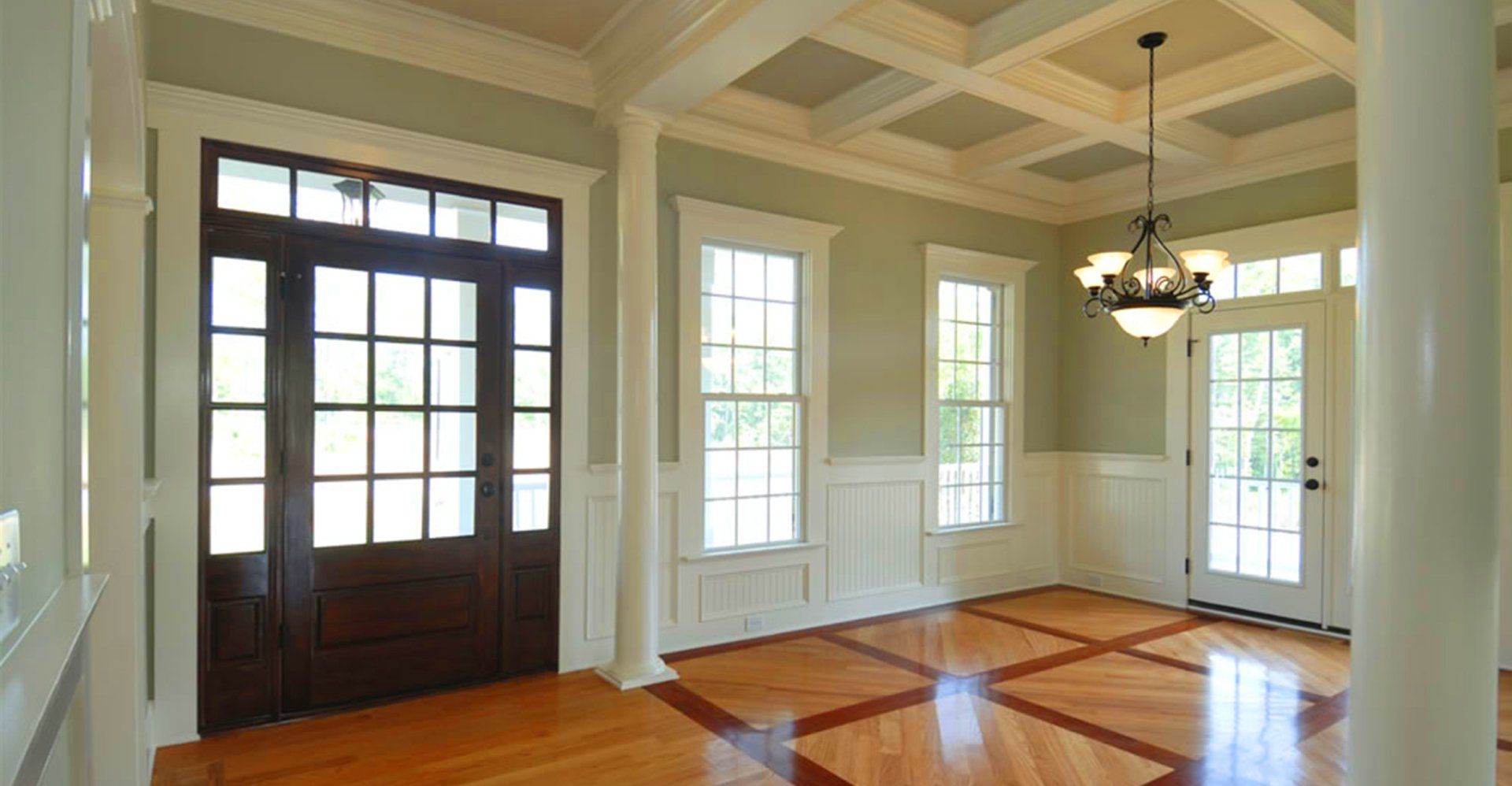 Elegant entryway with dark wood door, windows, columns, light wood floors, and coffered ceiling.
