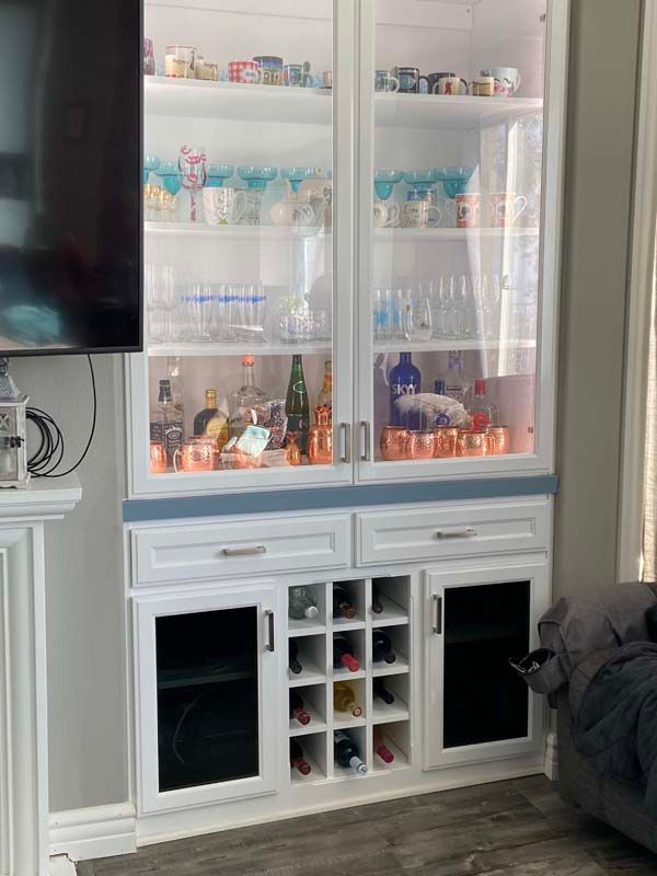 White built-in bar cabinet with glass doors, wine rack, and a collection of glassware and liquor.