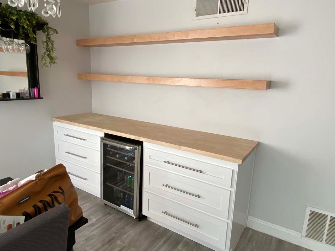 Built-in home bar with a wine fridge, white cabinets, light wood shelves, and countertop against a gray wall.