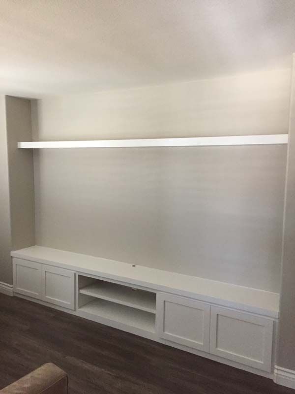 White built-in media center with a shelf above it, against a light gray wall. Wooden floors.
