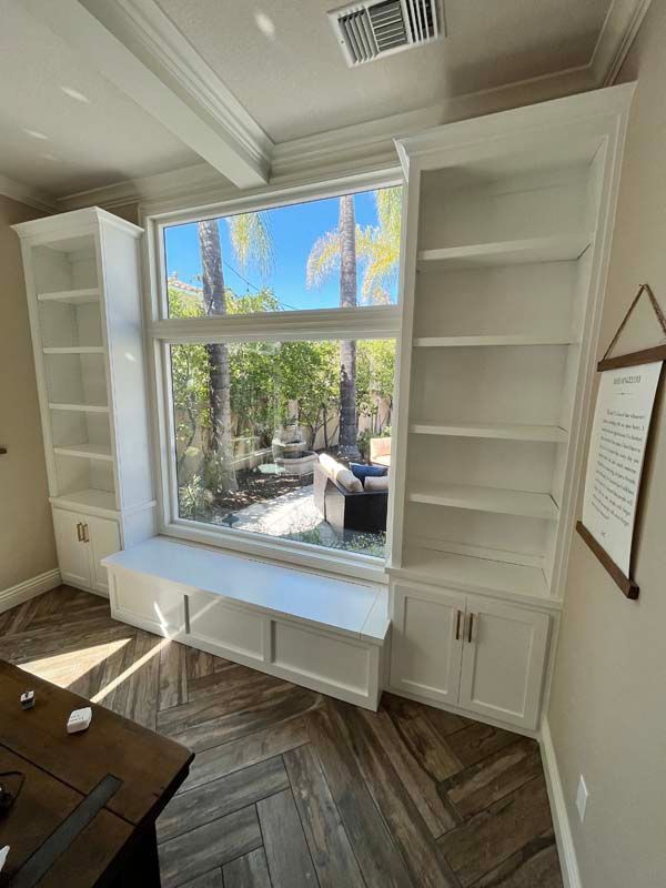 Built-in white bookshelves flank a large window with a seating area. The window overlooks a yard, the floor is herringbone.