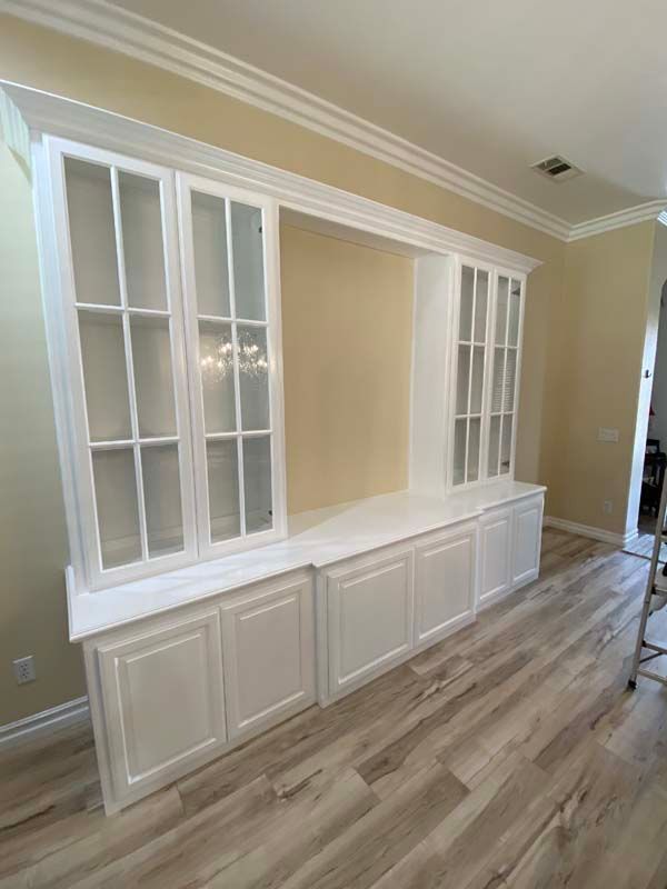 White built-in cabinet with glass doors and base cabinets, set against a beige wall, with wood flooring.