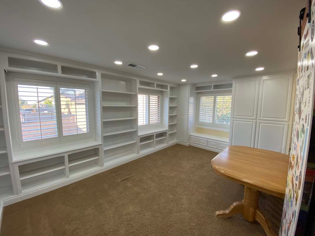 White-walled room with built-in bookshelves, windows, and a round wooden table on a brown carpet.