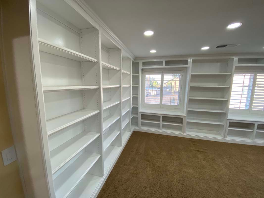 White built-in bookshelves in a room with brown carpet. The shelves surround a window and are empty.