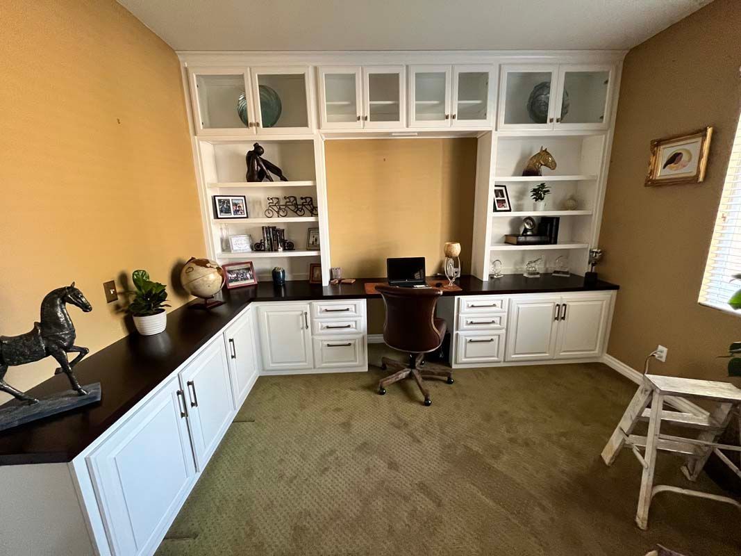 Built-in white desk with upper cabinets, shelves, and a workspace. Dark wood counter tops, office chair, and decor on shelves.