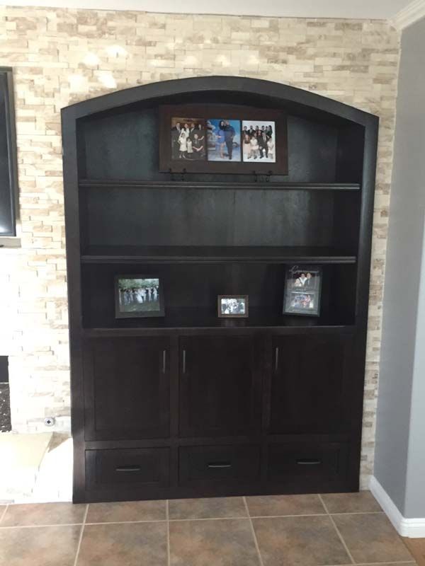Dark brown wooden bookcase against a brick wall, with arched top, shelves, cabinets, and drawers.