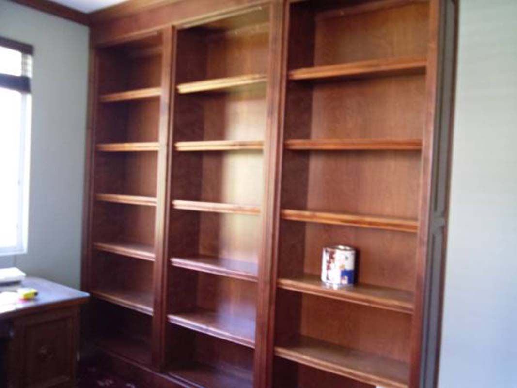 Built-in wooden bookshelves, three sections, stained brown, against a wall. One shelf holds a paint can.